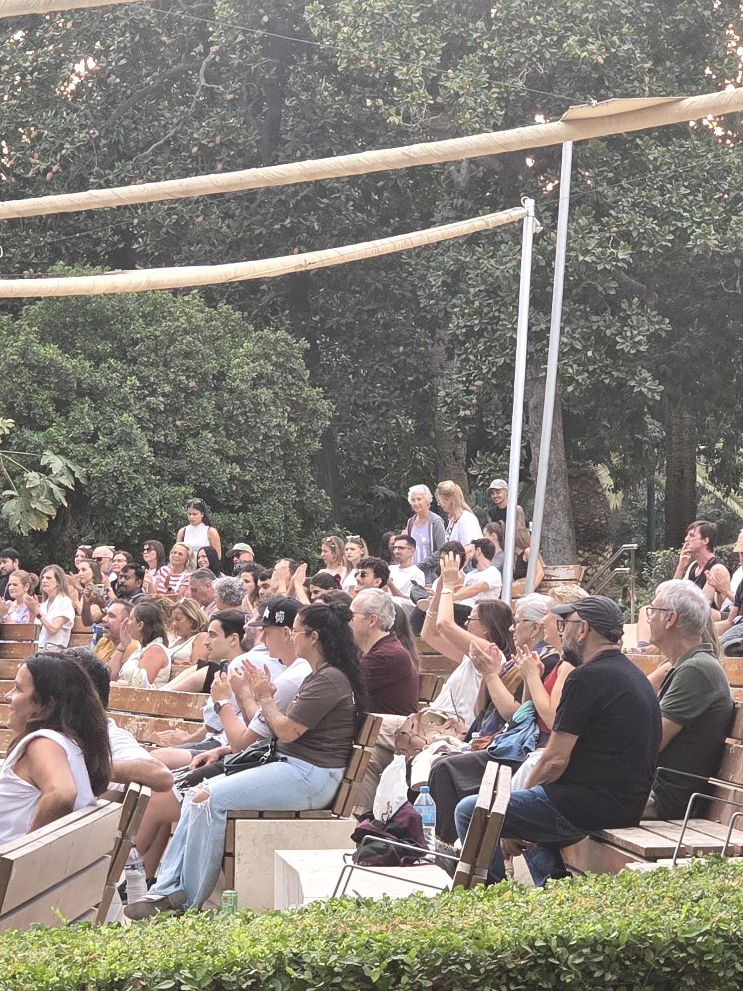 The image depicts an outdoor audience seated on tiered wooden benches, facing an unseen stage or performance area. The setting appears to be a park or garden, with lush green trees and foliage forming the background. Two horizontal, cream-colored fabric-covered beams stretch across the top of the frame, supported by vertical poles. Several people are visible, dressed in casual clothing, with some appearing to clap or react to an event. Their ages range from young adults to older individuals. The lighting suggests it is daytime, possibly late afternoon. The presence of a specific type of seating and the overall ambiance suggest this could be an amphitheater or outdoor performance venue within a botanical garden or park in Málaga, Spain, such as the Jardines de la Finca de la Concepción or a similar public garden space known for hosting events.