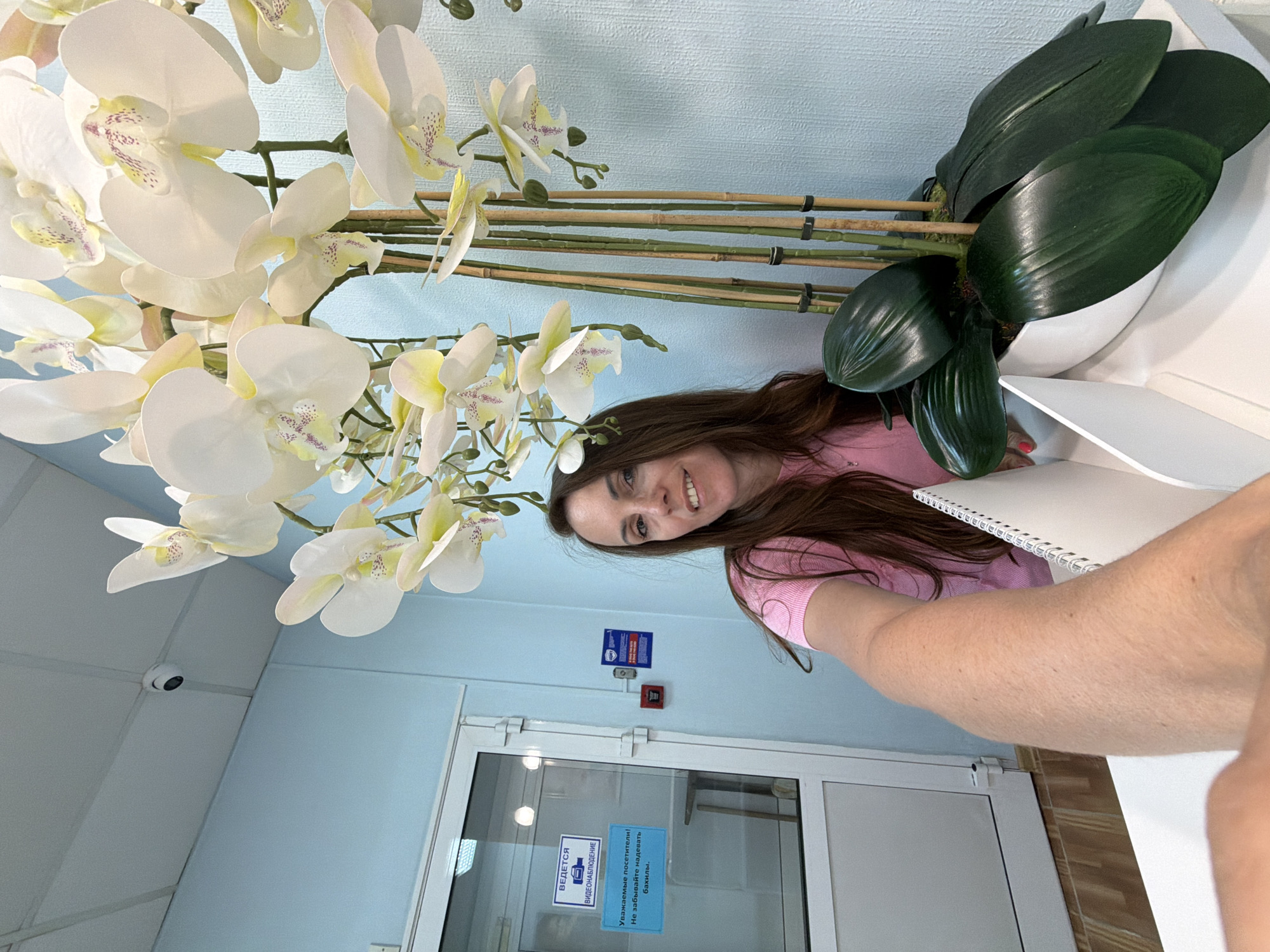 In Birobidzhan, Russia, a woman with long brown hair, wearing a pink top, smiles at the camera, holding up a notepad in a selfie-style shot. She is standing in front of a wall painted a light blue. To her left is a glass door with signs indicating "VIDEO SURVEILLANCE" and "Dear visitors! Please do not forget to wear shoe covers." A red fire alarm is mounted on the wall. To her right, a large artificial orchid plant with white flowers and green leaves in a white pot stands on a white counter. A security camera is visible on the white ceiling.