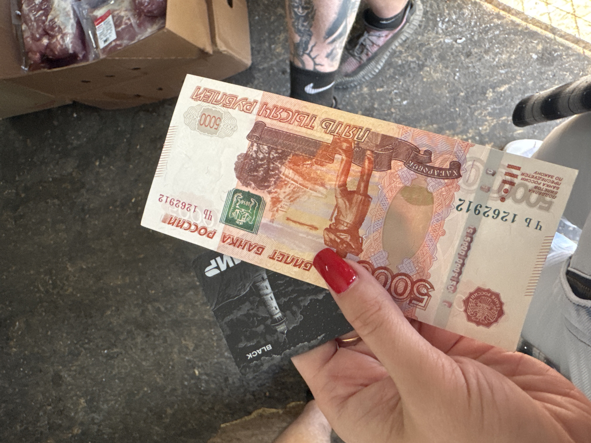A close-up, high-angle, outdoor, daylight shot shows a person's hand holding Russian currency and a black card. The hand, with red nail polish and a ring, is in the foreground. Behind it, a 5000 ruble banknote is visible, featuring text in Russian and an image of a monument. A black card, with "BLACK" printed on it and an image of a lighthouse, is partially obscured by the banknote. In the background, a cardboard box filled with packaged meat and a concrete surface are visible. The scene appears to be in an urban or industrial setting.