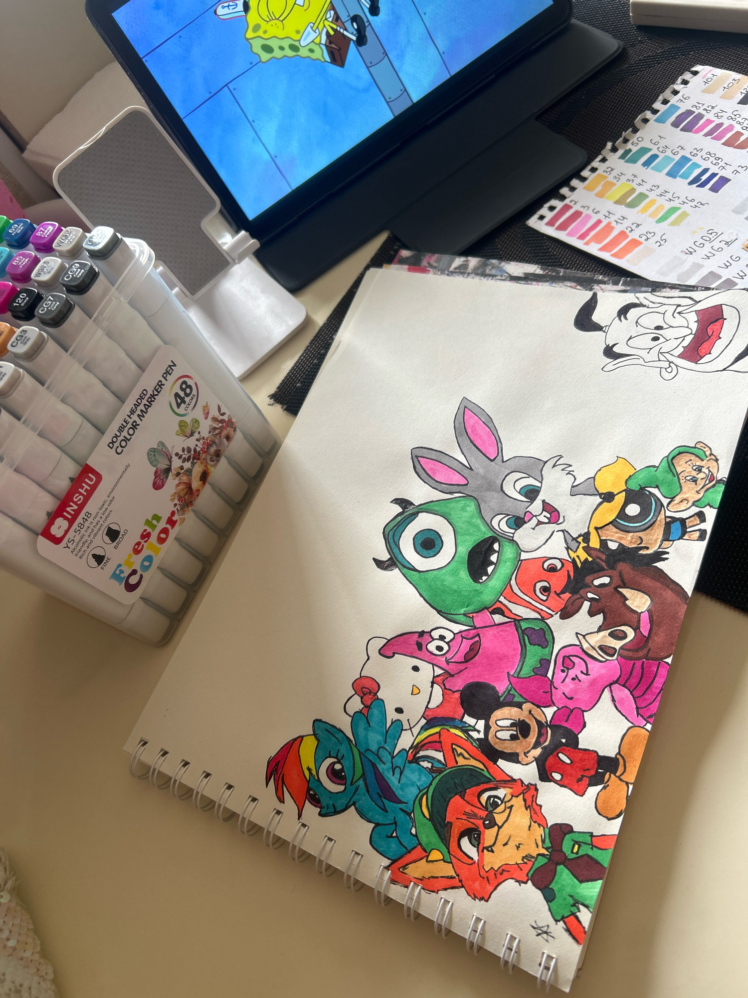 The image displays a close-up, high-angle view of a desk with various art supplies and a drawing. A tablet, displaying a cartoon image of SpongeBob SquarePants, is propped up on a stand. To the left, a clear plastic container filled with dual-tipped color markers is visible. A colorful swatch chart for markers is situated to the right of the tablet. In the foreground, a spiral-bound sketchbook lies open, showcasing a vibrant drawing composed of multiple cartoon characters, including Hello Kitty, Mickey Mouse, Rainbow Dash, and characters from Looney Tunes and The Powerpuff Girls. The drawing is rendered in colored markers. The background consists of a dark textured surface, possibly a mousepad. The overall scene appears to be an indoor setting, likely a home or studio, with no discernible elements that would specifically place it in the city of Khyrdalan, Azerbaijan.