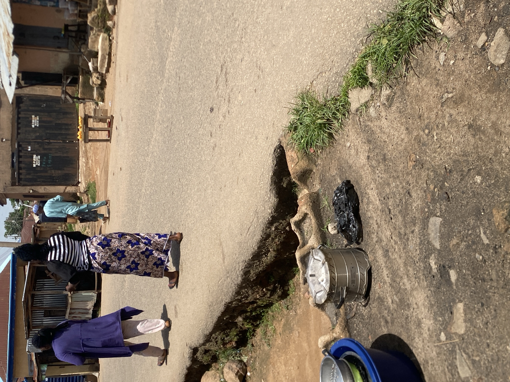 Two women are walking on a paved road in Jos, Nigeria, heading away from the camera. The woman on the left wears a purple tunic over white trousers and sandals. The woman on the right wears a black and white striped top and a long blue and white floral skirt, also with sandals. In the foreground, on the left, a blue basin contains a metal colander and a green item, possibly food. Next to the basin is a silver, cylindrical charcoal stove. To the right of the stove is a black plastic bag, appearing to contain charcoal. A man in a light blue robe and a blue cap is visible in the background, walking in the same direction as the women, carrying a dark bag. The environment appears to be a street with buildings and shops in the background. The road shows signs of wear and tear, with some debris and vegetation growing along the edges.