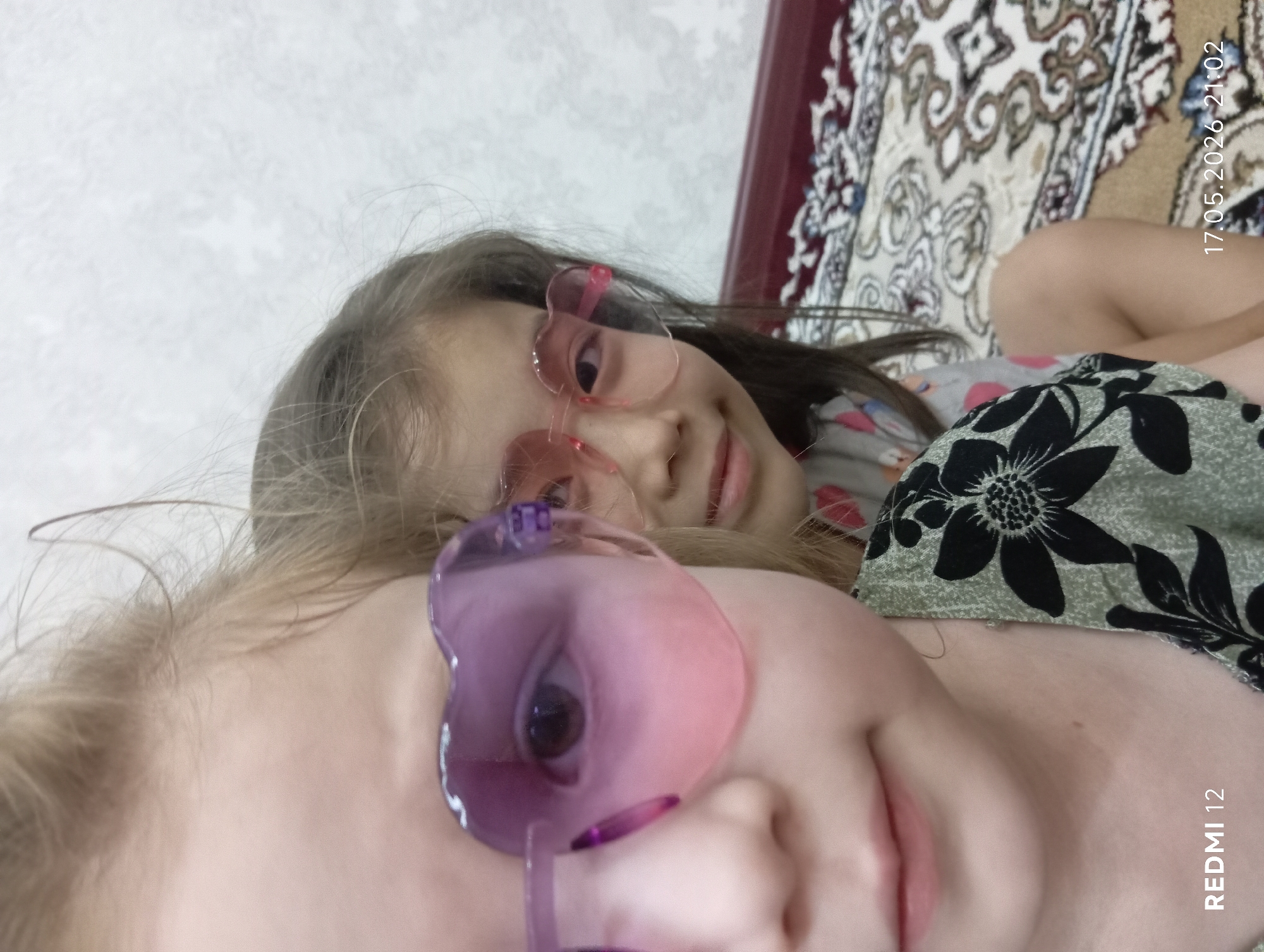 The image shows two girls wearing heart-shaped glasses. One girl is lying on top of the other. The top girl has blonde hair and is wearing purple glasses. The other girl has brown hair and is wearing pink glasses. They are lying on a patterned blanket near a wall. The date "17.05.2026 21:02" is visible in the corner of the image along with the text "REDMI 12". There is no information in the image about the city of Bekabad, Uzbekistan.