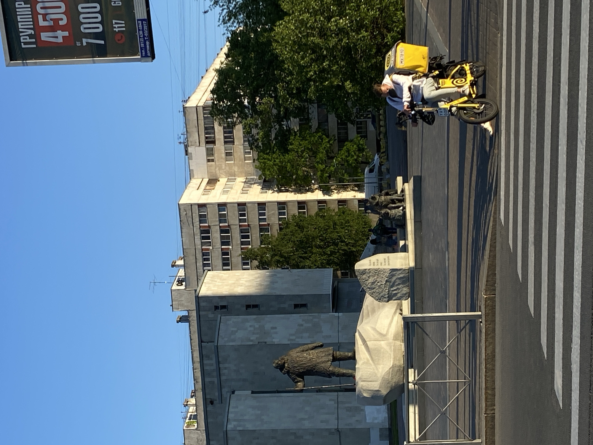 The image shows a monument to Lenin in Saint Petersburg, Russia. The monument is located on Moskovsky Prospekt, and it depicts Lenin in a long coat, standing on a pedestal. In the background, there is a multi-story building, and a blue sky is visible above the building. A delivery driver on a yellow bike with a yellow backpack is shown at the crosswalk. A billboard is seen with Russian text on the left of the image. The ground is paved with asphalt, and there are white stripes on the crosswalk.