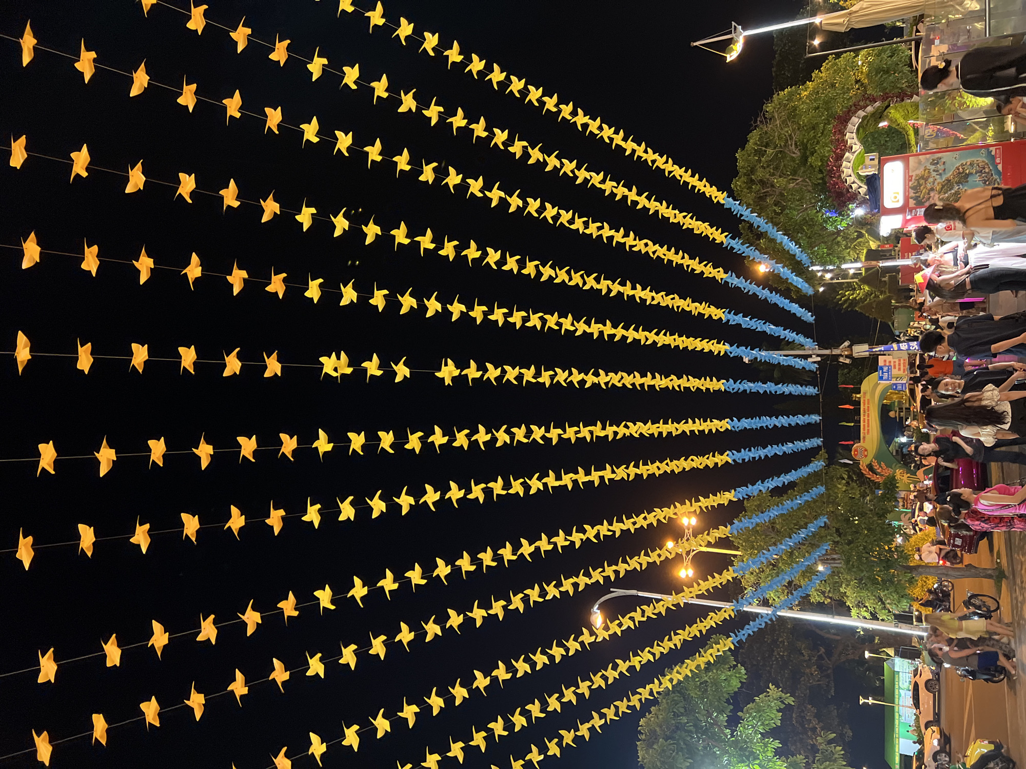 The image depicts a nighttime scene in Nha Trang, Vietnam, likely during a festival or event. Overhead, strings of yellow and blue paper pinwheels are suspended against a dark sky, creating a festive ambiance. Below the pinwheels, people are gathered on a sidewalk, illuminated by streetlights and decorative lighting. Several cars and motorcycles are visible, suggesting a public thoroughfare or plaza. In the background, a large archway with decorative elements and banners indicates a focal point for the event. The overall atmosphere is lively and celebratory, characteristic of a night market or cultural gathering in a Vietnamese city.