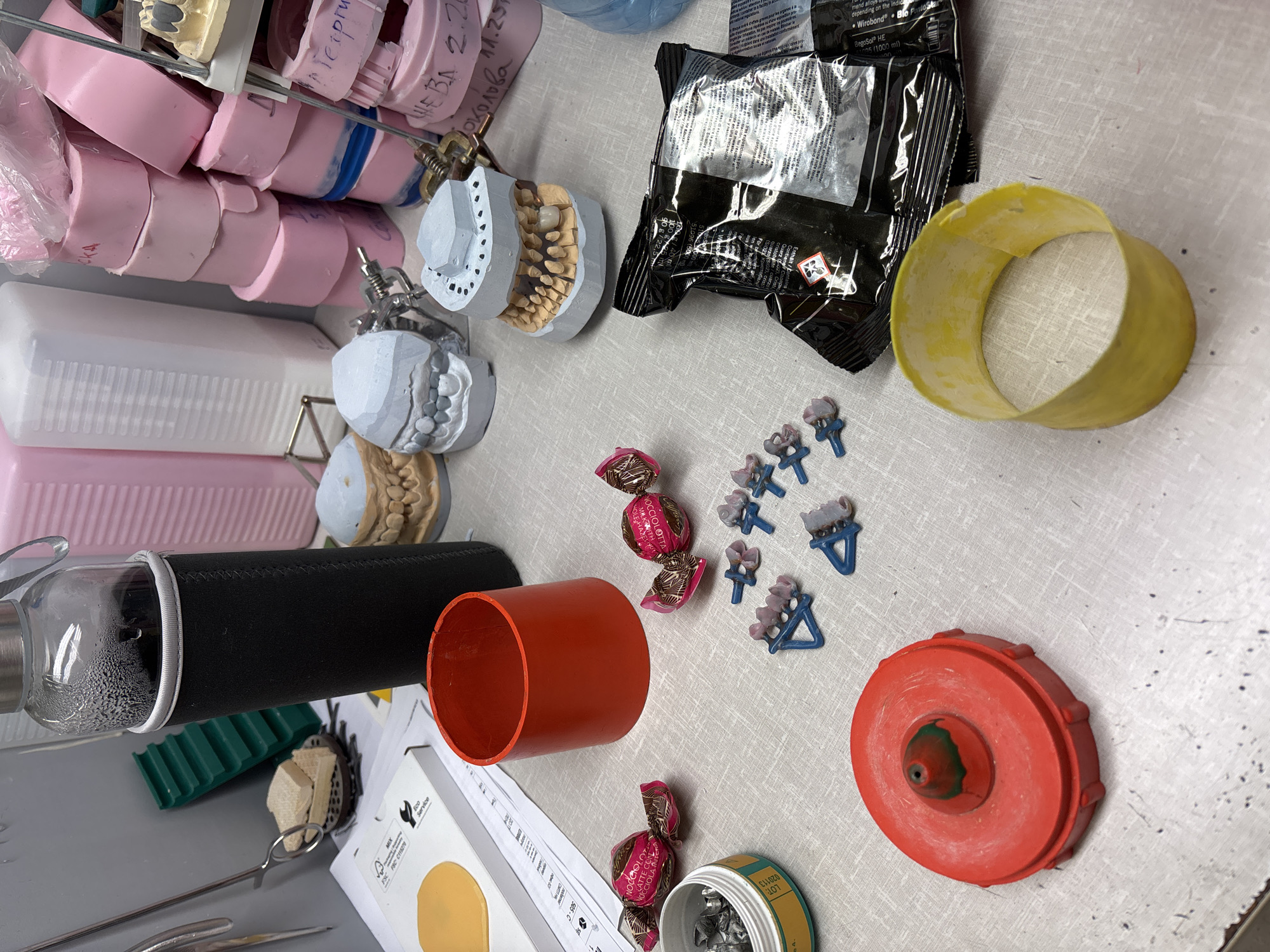 The image shows a table surface covered with various dental and crafting materials. There are numerous pink and white dental molds, as well as dental casts with articulated teeth. Several pieces of dental equipment are present, including articulated mounts. There is also an opened black packet of dental materials with hazard symbols and text in English. A yellow mold and several small blue dental casting fixtures are visible. Additionally, there are several wrapped chocolate candies, an open container of dental materials, a red cylinder, a glass water bottle in a black sleeve, and other tools and documents. The location appears to be a dental laboratory or workshop. There is no information to indicate the city of Sofia, Bulgaria is correct.