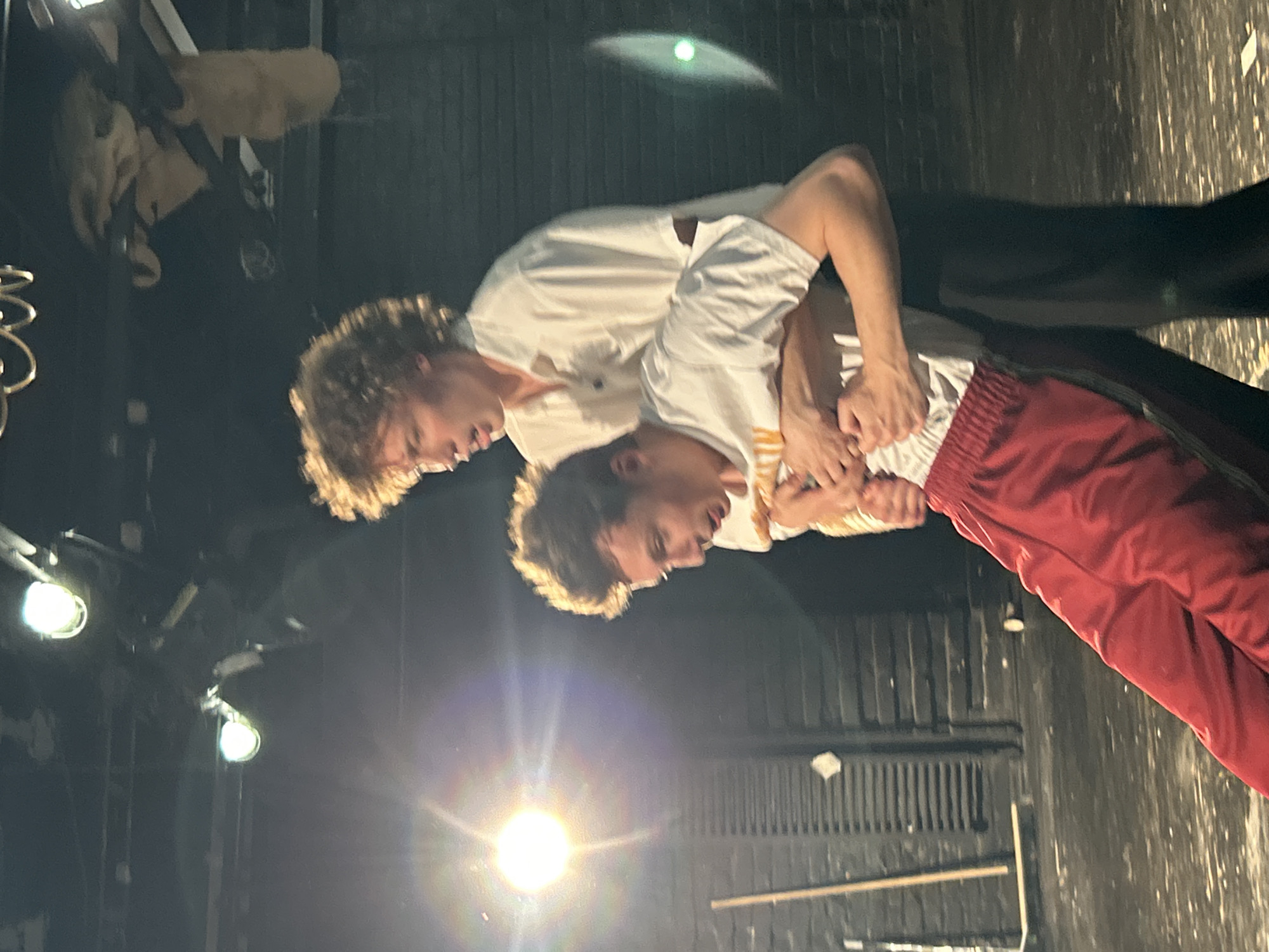 Two young men are engaged in a theatrical performance on a stage. The man in the foreground, wearing red athletic pants and a white t-shirt with yellow markings, is being held from behind by the man standing behind him. The man in the background is wearing a white button-down shirt and dark pants, and his curly hair is illuminated by stage lighting. The stage appears to be a dark, indoor space with exposed lighting fixtures and a brick wall in the background. The lighting is dramatic, with bright spots and shadows. The overall impression is one of intense physical interaction, likely part of a dramatic scene. The image was taken at an unknown location in Unknown, Unknown.