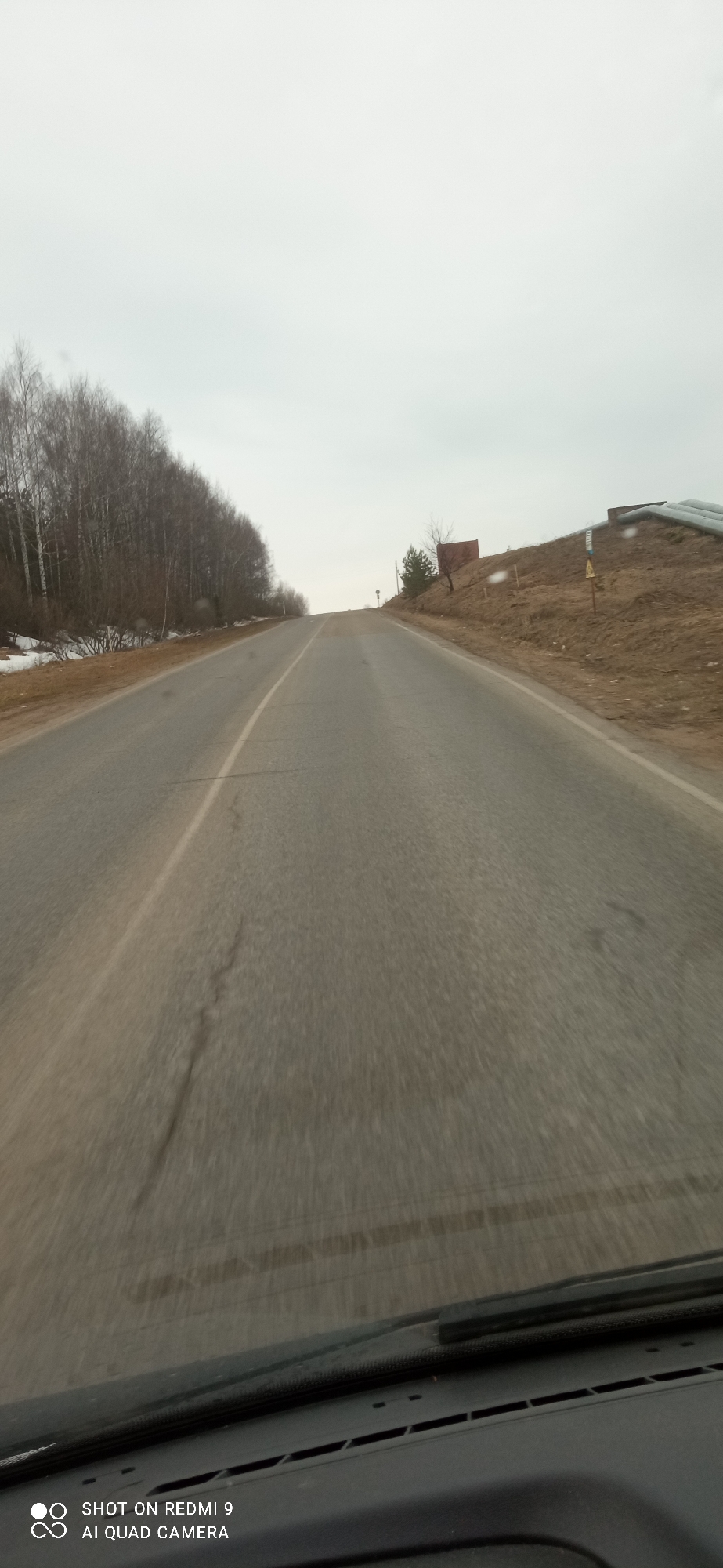 The image shows a view from inside a vehicle traveling on a gray asphalt road that ascends a slight incline. A solid white line marks the road's center. To the left of the road, a line of bare trees stands, with a patch of snow visible near the ground. On the right, the ground rises, revealing an embankment with a small building and some pipes. The sky is overcast. In the foreground, the vehicle's dashboard and windshield wiper are visible. The image was taken in Ольховка, Russia.