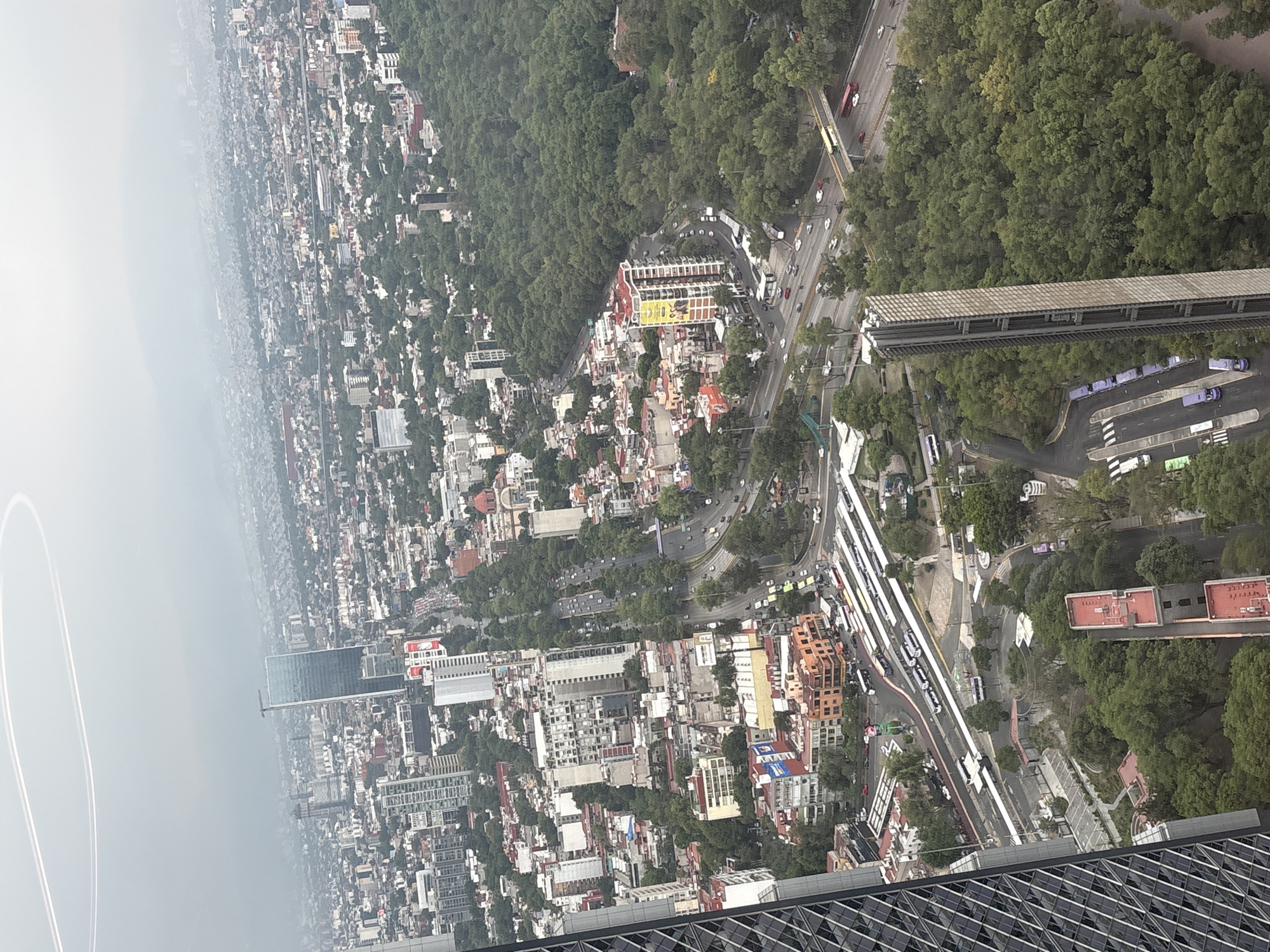 The aerial shot depicts a densely populated area in Mexico City, Mexico, with a mix of residential and commercial buildings interspersed with trees and green spaces. In the foreground is the edge of a modern high-rise building, with a geometric patterned facade. Streets are visible, carrying vehicular traffic. Adjacent to the built environment lies a forested area that appears to be Chapultepec Park. In the distance, the urban sprawl extends into a hazy horizon.