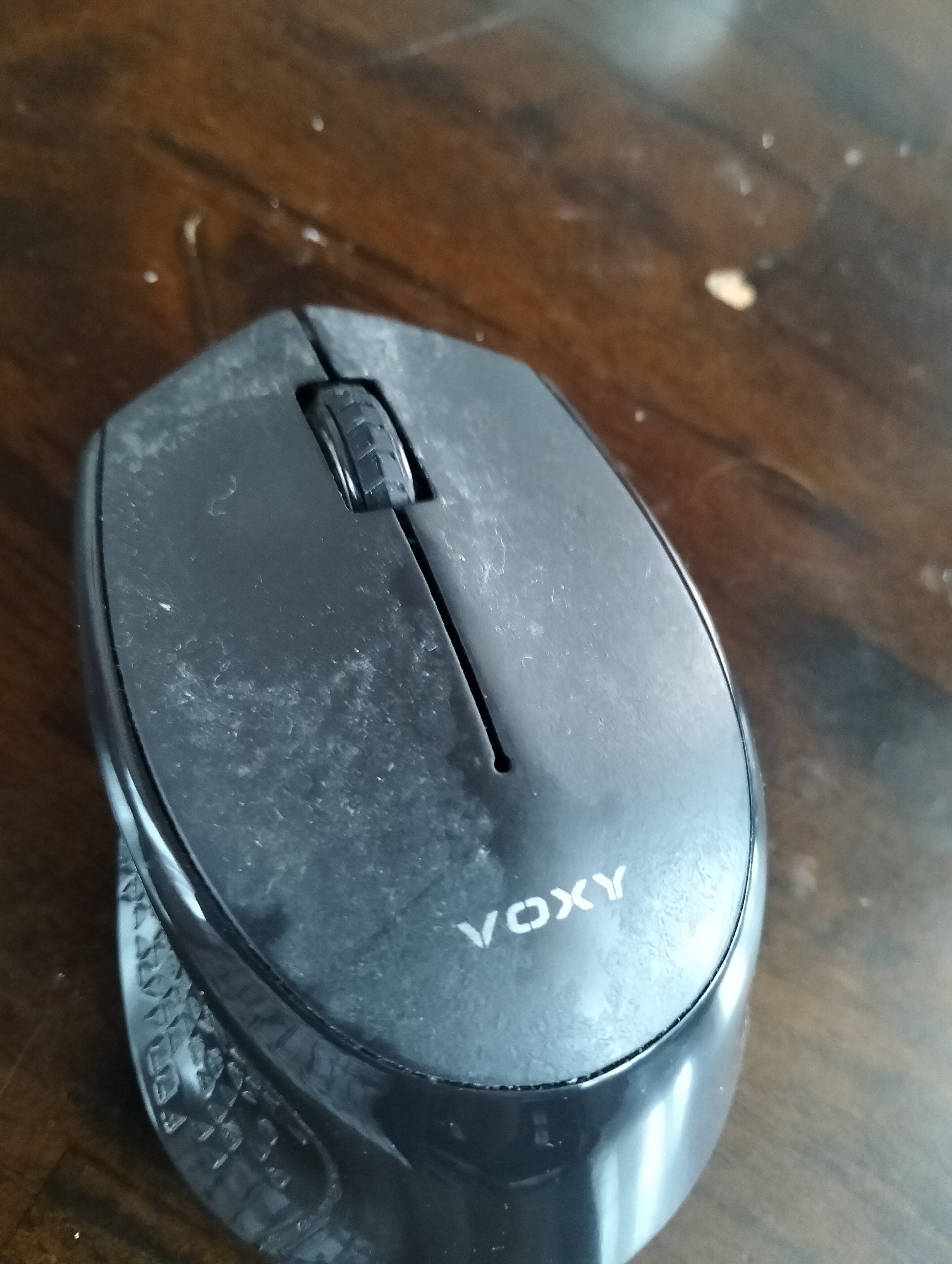 A dusty, black wireless computer mouse with the brand "VOXY" visible on its surface sits on a wooden table. The mouse has a scroll wheel in the center and a textured grip on the side. The lighting suggests an indoor environment, possibly a home or office. There is no indication of the city of Julicotmeurak, Indonesia, or any specific actions or interactions taking place.