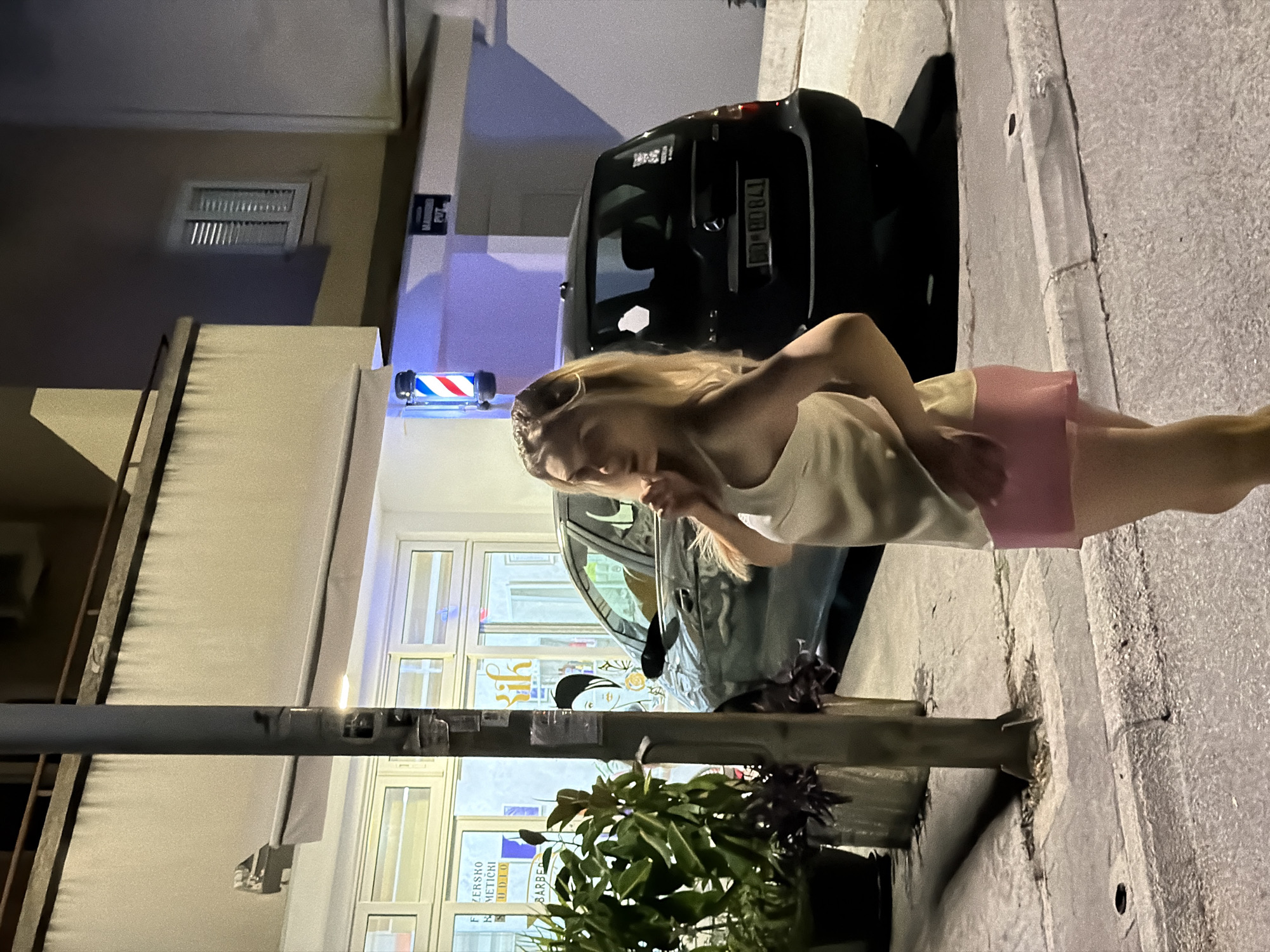 A blonde woman stands on a sidewalk at night in front of a storefront in Budva, Montenegro. She is wearing a light-colored tank top and pink shorts. She has her hand near her mouth and is looking to the left. Behind her, a dark-colored car is parked on the street. The storefront appears to be a barber shop, indicated by a barber pole with red, white, and blue stripes, and signage in a foreign language. A street light is visible on the left side of the image. The ground is paved with asphalt and concrete.