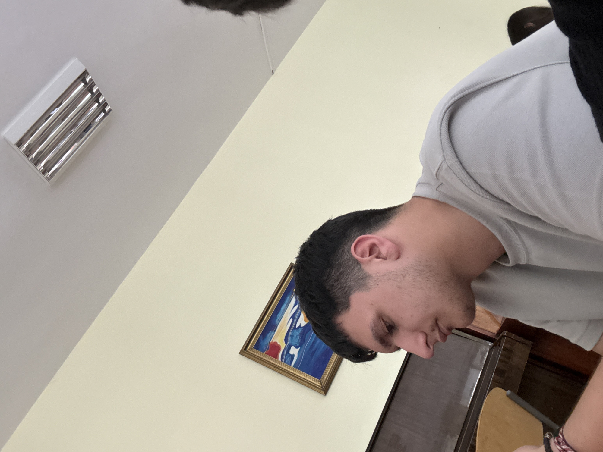 A young man, seen in profile from the right side, is the main subject of the image. He has short, dark hair and light stubble, and is wearing a light grey, short-sleeved collared shirt. His head is tilted downwards, indicating he is looking at something out of frame below him, seemingly focused or reflective. A colorful, woven bracelet adorns his right wrist.
The setting is indoors. A white ceiling with a rectangular fluorescent light fixture occupies the upper left portion of the image. Below the ceiling, a pale yellow wall covers much of the background. Hanging on this wall is a framed painting with a golden, ornate frame. The artwork within features abstract shapes primarily in blue and yellow, with some red accents. In the lower right, part of a dark wooden furniture item, possibly a cabinet or desk, is visible, along with a light-colored wooden chair back.
The scene depicts a quiet indoor moment, possibly in a study, office, or classroom setting. No specific time of day is discernible, as the lighting appears to be artificial. There is no visible text in the image. The location context for this scene is Kardzhali, Bulgaria.
