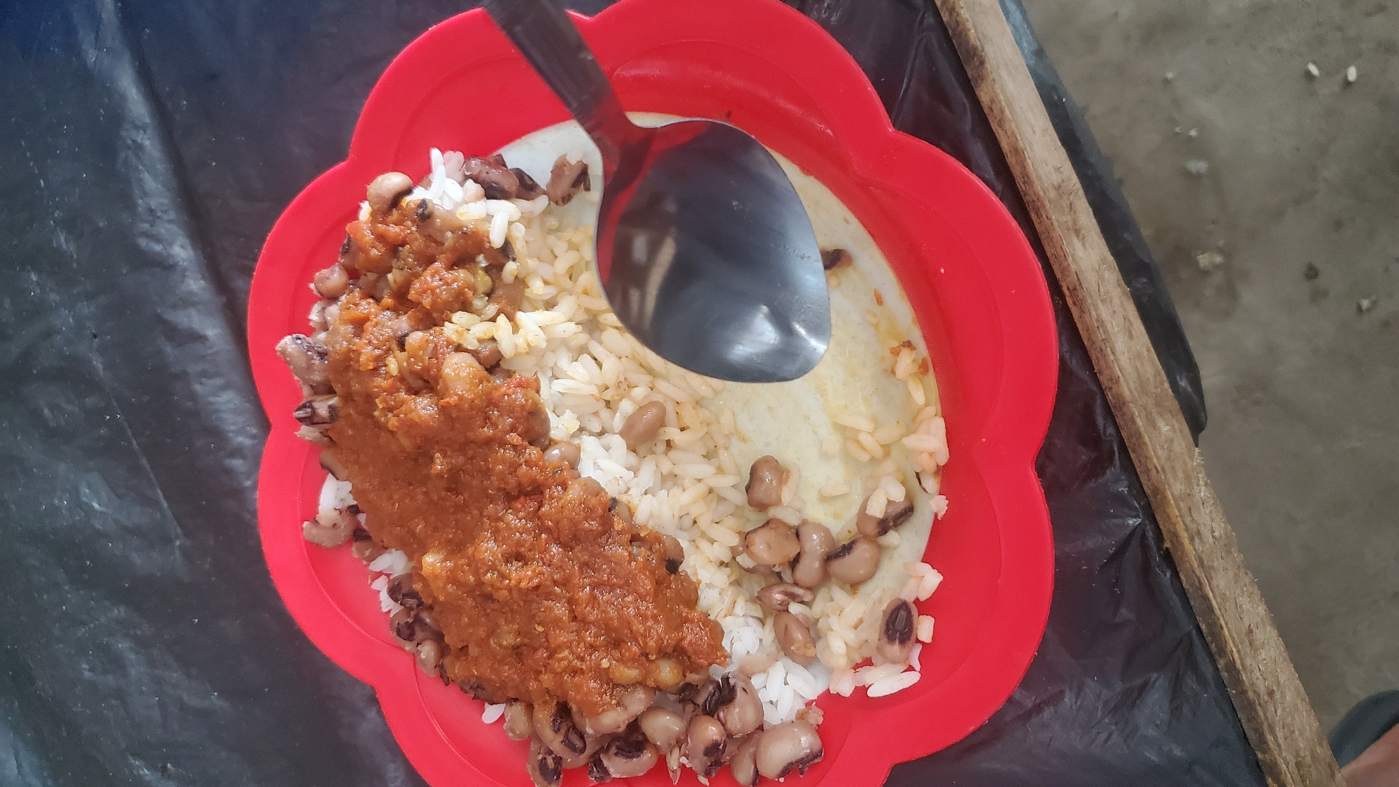 A close-up, overhead shot shows a red, scalloped plate filled with rice, black-eyed peas, and a reddish-brown sauce. A silver spoon rests on the right side of the plate, partially submerged in the rice. The food is served on a black, textured surface, possibly plastic sheeting, which is laid on top of a wooden surface. The background is dark and out of focus. The image was taken in Jalingo, Nigeria.