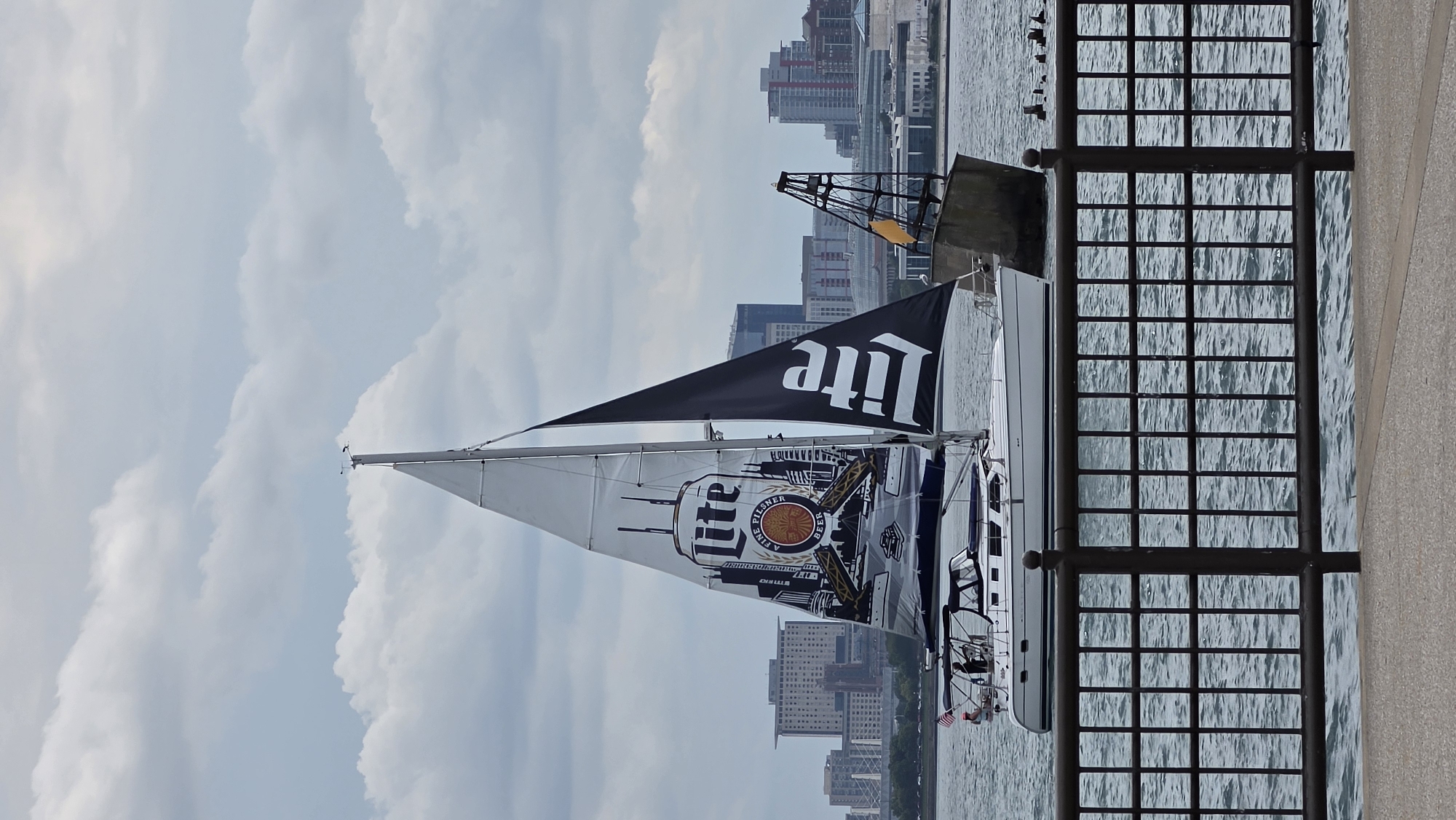 A sailboat with a large "Lite" beer logo printed on its mainsail sails on the water with a city skyline in the background. The boat is white with a dark blue canopy over the cockpit. The mainsail is white with a graphic of a Lite beer can and city buildings. A black jib sail also has the "Lite" logo. In the background, a dense collection of modern buildings and skyscrapers form the Chicago skyline. The water appears choppy, and the sky is overcast with clouds. In the foreground, a dark metal fence runs horizontally across the image, and a paved walkway is visible at the very bottom. A navigational buoy or marker is visible to the right of the sailboat.