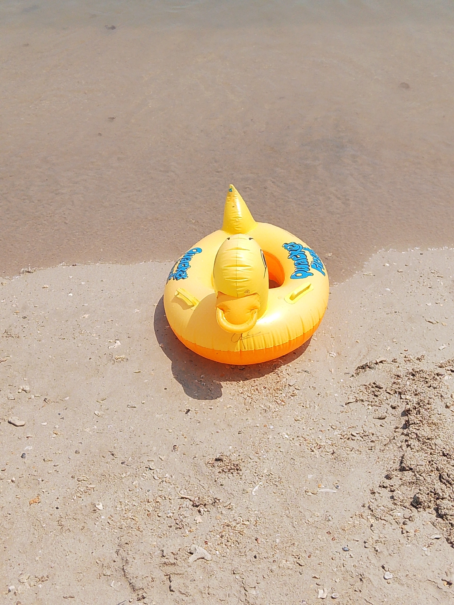 A bright yellow inflatable duck with blue lettering, labeled "Buddy", rests on a sandy beach next to the shallow, clear water of a body of water. The duck is positioned in the sand, with its rear end slightly submerged in the water. The water's edge creates a distinct line across the lower portion of the image, with fine sand and small shell fragments scattered on the beach. The sky is not visible, but the bright sunlight casts a shadow from the inflatable duck onto the sand. The scene appears to be a tranquil beach setting in Pejagan, Indonesia.