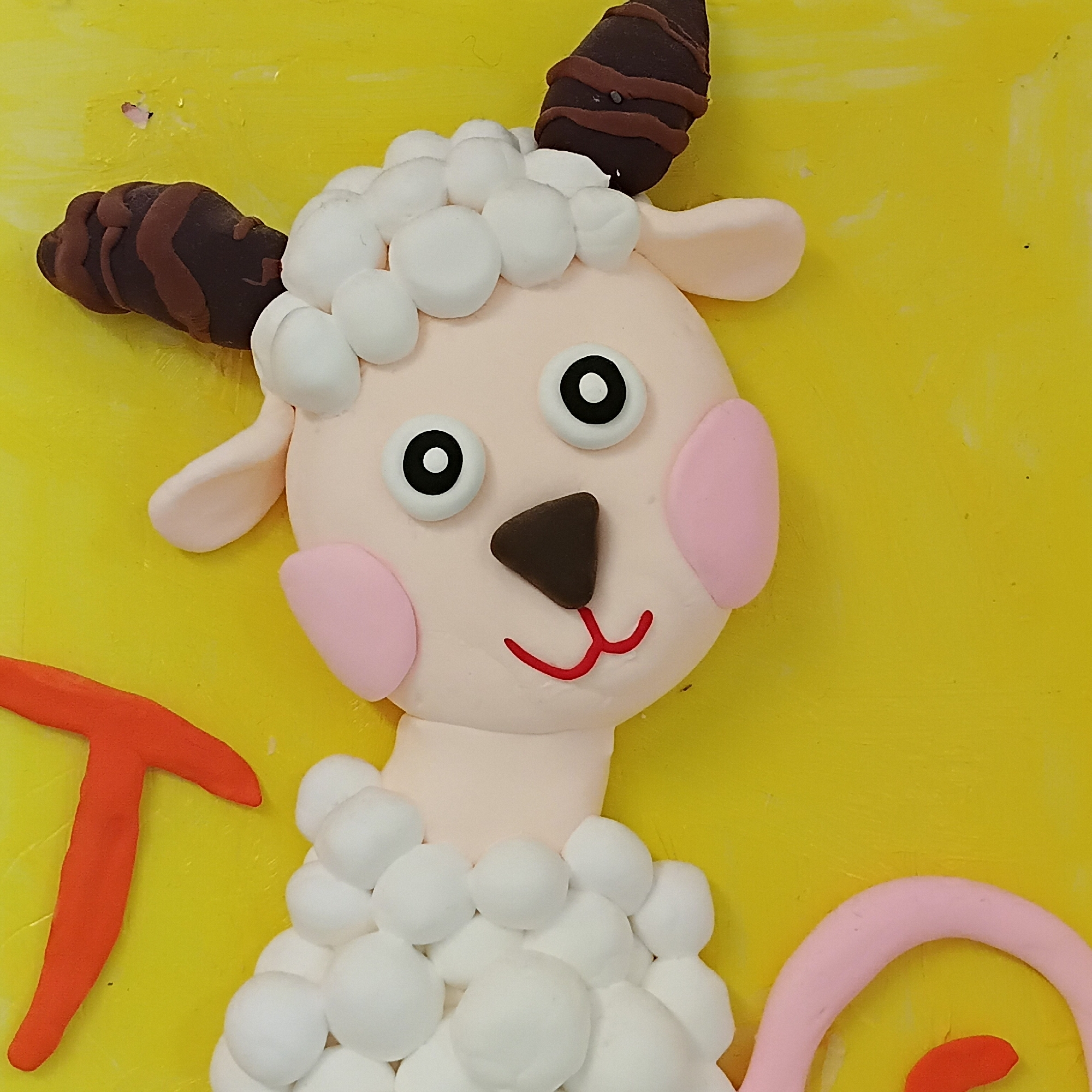 This is a close-up shot of a vibrant, three-dimensional craft artwork, likely made from modeling clay or play-doh, originating from Shunde District, China. The central subject is a charming, cartoon-style sheep or goat character. Its face and neck are light pink, featuring large, round white eyes with black pupils, a dark brown triangular nose, and a cheerful red upturned mouth. Oval-shaped pink blush marks adorn its cheeks.
The creature's wool is represented by numerous small, white, spherical shapes, giving it a fluffy appearance around its head and body. Two dark brown, ridged horns with lighter brown stripes protrude from the top of its head.
The background is a bright, textured yellow, possibly a painted surface or another layer of modeling material. To the left, part of a bold orange letter 'T' is visible. On the right, a curved pink shape, possibly a tail or another decorative element, extends into the frame. The overall aesthetic is childlike and whimsical, characteristic of a craft project designed for or by children.