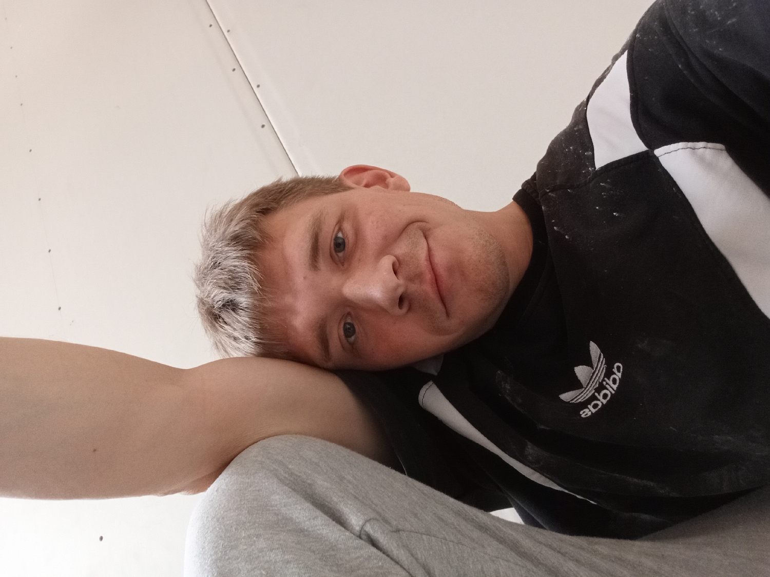A man is depicted in a low-angle, close-up shot, likely a selfie, looking directly at the camera with a slight, relaxed smile. His head is tilted and resting on his bent left arm, while his body extends out of frame. He has short, light brown hair with notable lighter or greyish strands at the front, and light-colored eyes. Stubble is visible on his chin and jawline.
He wears a black long-sleeved shirt or sweatshirt featuring white stripes on the sleeves and a white Adidas trefoil logo on the chest. The logo appears reversed, reading "sabbida," suggesting a front-facing camera or mirrored image. The shirt is visibly speckled with white particles, consistent with dust, paint, or plaster, indicating possible involvement in construction or renovation activities. A section of grey fabric, likely his trousers, is visible at the bottom of the frame.
The background consists of a plain, light-colored surface, possibly a wall or ceiling. Small, dark, regularly spaced dots, which appear to be screw heads or nails, are visible on this surface, suggesting an unfinished interior or a space undergoing construction in Chaikovsky, Russia. The overall impression is one of casualness and perhaps a brief break from work.