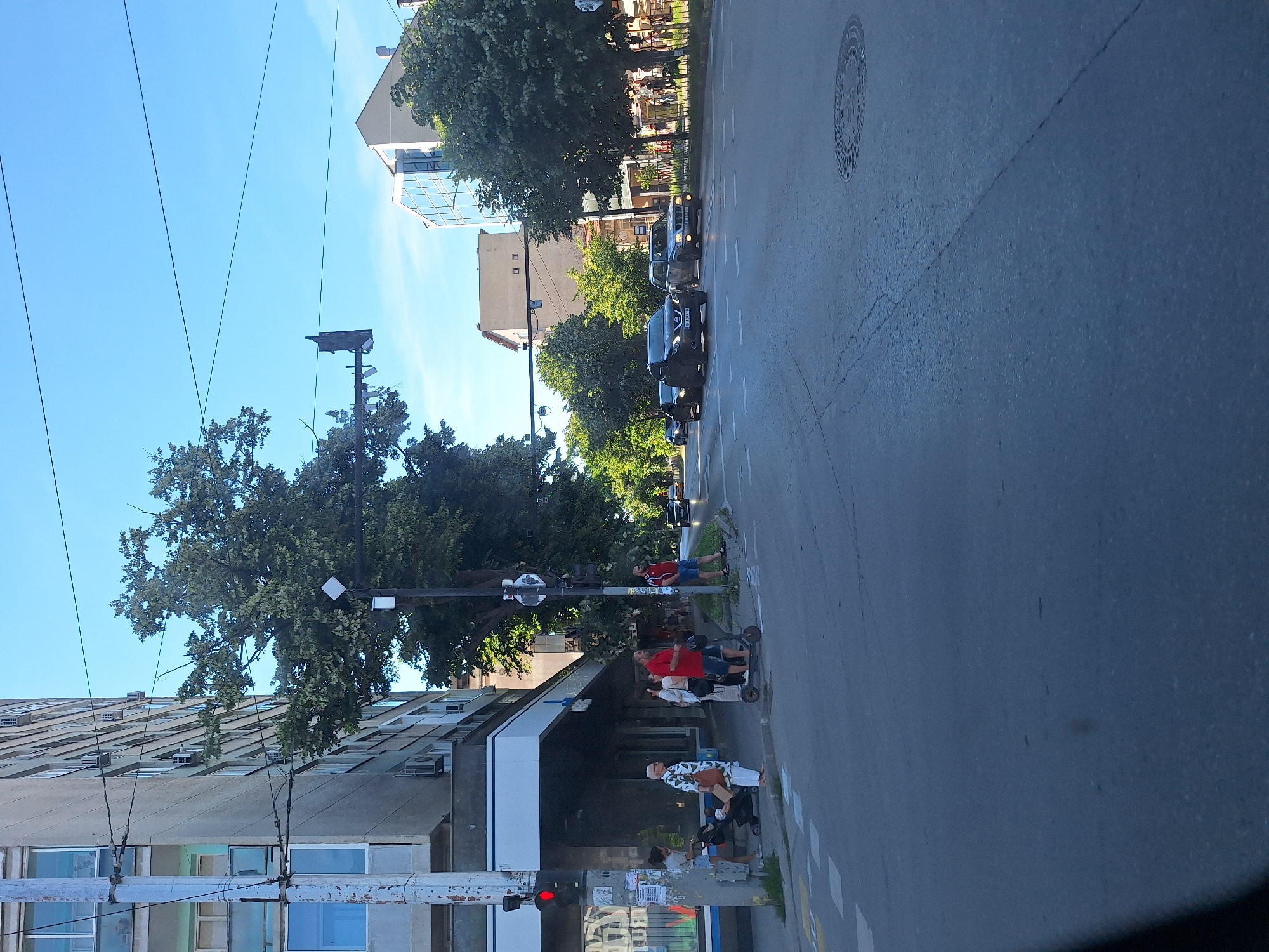 The image depicts a street scene in Varna, Bulgaria, under a clear blue sky, suggesting a sunny day. The perspective is from inside a vehicle, looking outwards.
The foreground features a concrete building with multiple windows and air conditioning units. Electrical wires are visible crisscrossing the frame. A traffic light shows a red signal.
Further down the street, several people are visible, some standing and others on what appear to be scooters. One person in a red shirt and blue shorts is standing near a scooter, while another person in a patterned shirt and white pants is seated on a scooter. The presence of these individuals suggests everyday life and pedestrian activity.
A line of cars is parked or moving along the street. Behind the cars, lush green trees line the sidewalk, providing shade and natural beauty to the urban environment. Buildings of varying architectural styles can be seen, including a modern building with a glass facade. A circular manhole cover is visible on the road surface.
The overall impression is a typical urban street scene during daytime, with a mix of residential and commercial elements, and people engaged in their daily routines. There is no visible text in the image that provides specific information.