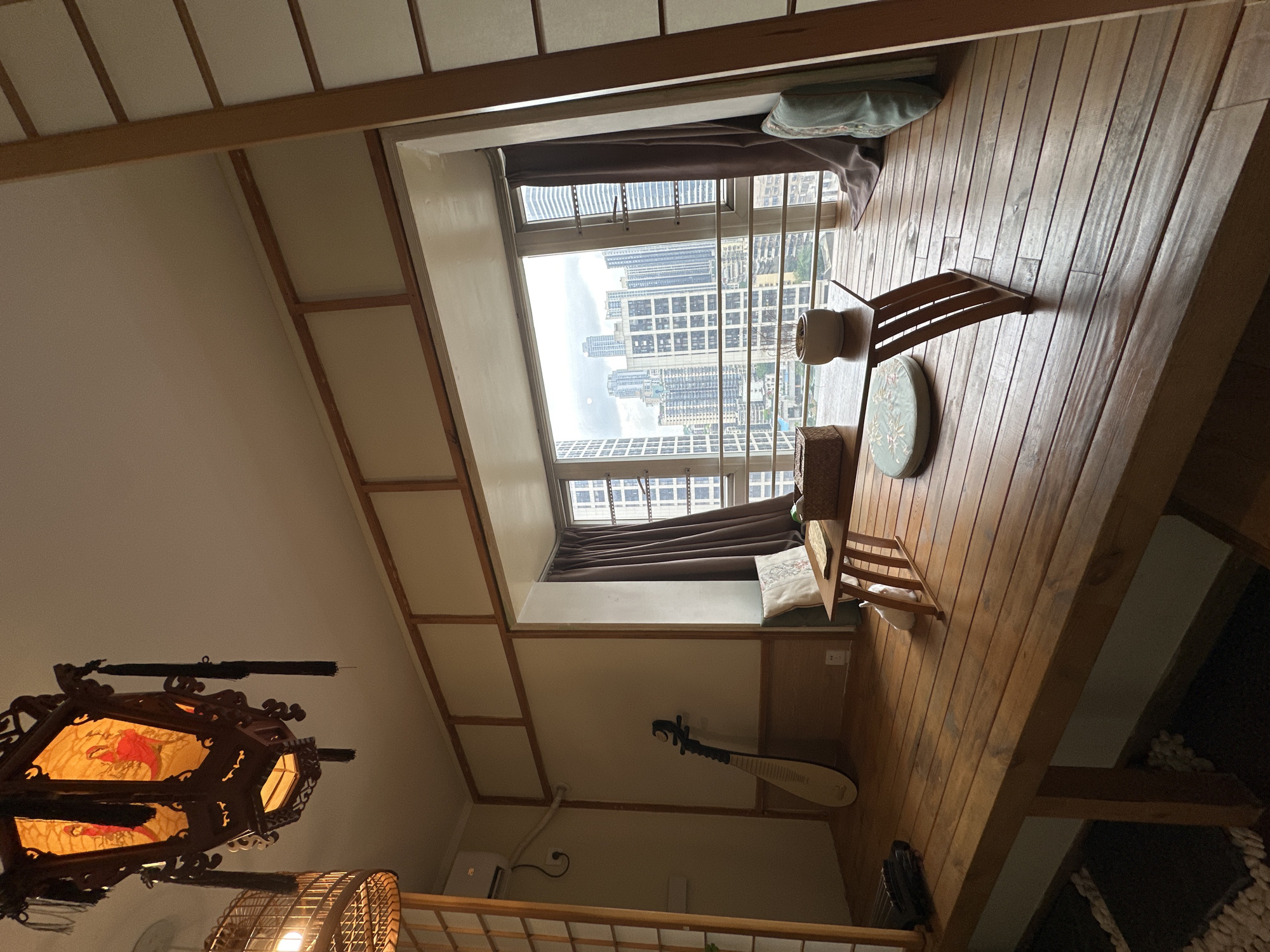 The image depicts an interior space with a unique architectural design. The room features a raised wooden platform with a window offering a view of a dense urban landscape, presumably in Yuzhong District, China, given the context. High-rise buildings dominate the view. A small table and chairs sit on the wooden platform. A traditional Chinese musical instrument, likely a Pipa, leans against the wall. Two distinct light fixtures hang from the ceiling in the foreground. The lighting is relatively dim. There are no people visible in the image.