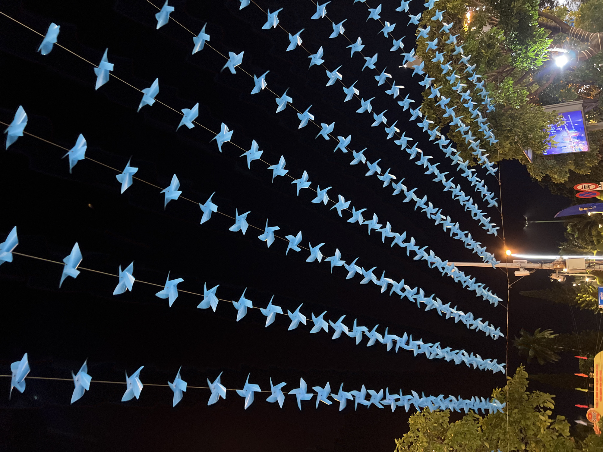 The image shows a street in Nha Trang, Vietnam, decorated with numerous blue origami birds strung together on thin wires, forming cascading lines against a dark night sky. These decorative strings are suspended overhead. In the lower portion of the image, trees with green foliage are visible, along with streetlights and utility poles. There are also glimpses of street signs, including one with Vietnamese text, and a large screen displaying colorful visuals. The overall scene suggests an outdoor evening event or festival.
