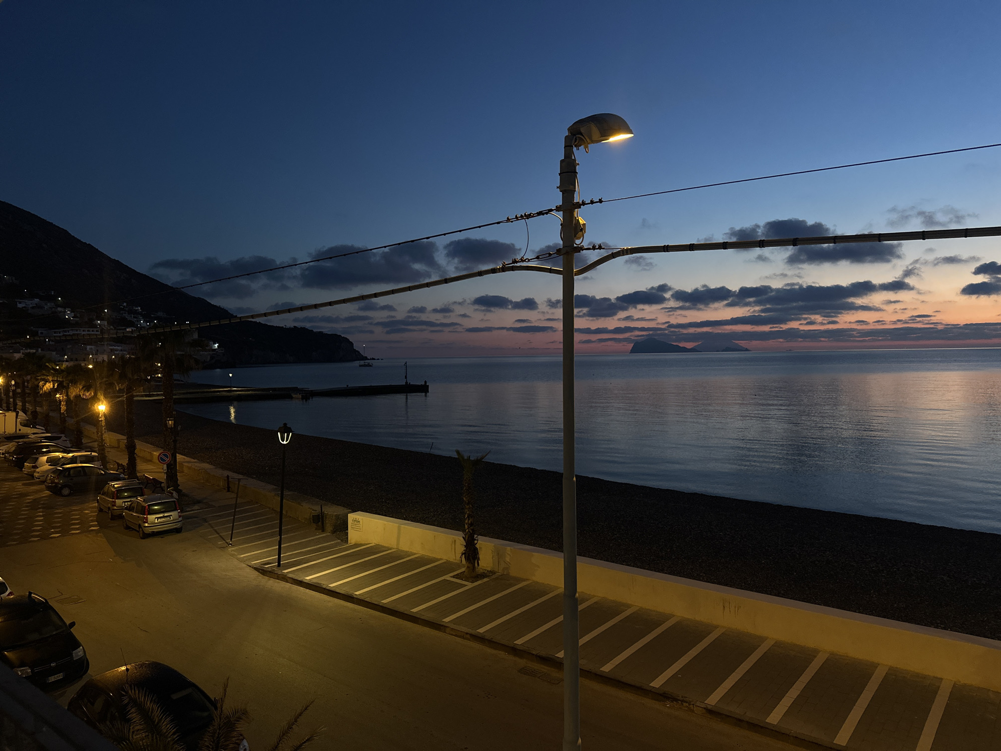 The image presents a tranquil twilight scene of a coastal town, identified as Lipari, Italy. The sky transitions from a deep, dark blue at the top to soft oranges and pinks at the horizon, indicating either sunrise or sunset. Wispy clouds are scattered across the sky, reflecting the fading light.
In the foreground, a paved road runs parallel to the coastline, illuminated by warm streetlights. Numerous cars are parked along the side of the road, and the yellow glow from the lights casts distinct shadows and highlights on the pavement and white parking lines. Several palm trees line the street, their dark silhouettes contrasting with the artificial light.
To the left, a steep, dark hillside rises, dotted with the clustered white buildings of a town. These structures show faint lights, suggesting habitation. A short pier or jetty extends into the calm, dark waters of the bay.
The central feature is a prominent metal streetlight with a bright, yellowish lamp, its light radiating downwards. Overhead cables are strung across the frame, attaching to the streetlight pole and extending horizontally, adding a linear element to the composition.
To the right, the calm sea stretches out, reflecting the twilight colors of the sky. A dark, pebble or shingle beach separates the road from the water. In the distant horizon, two small islands are silhouetted against the colorful sky. The overall atmosphere is peaceful and serene, with no human activity explicitly depicted, evoking a sense of quietude at the edge of the day. No discernible text is visible in the image.