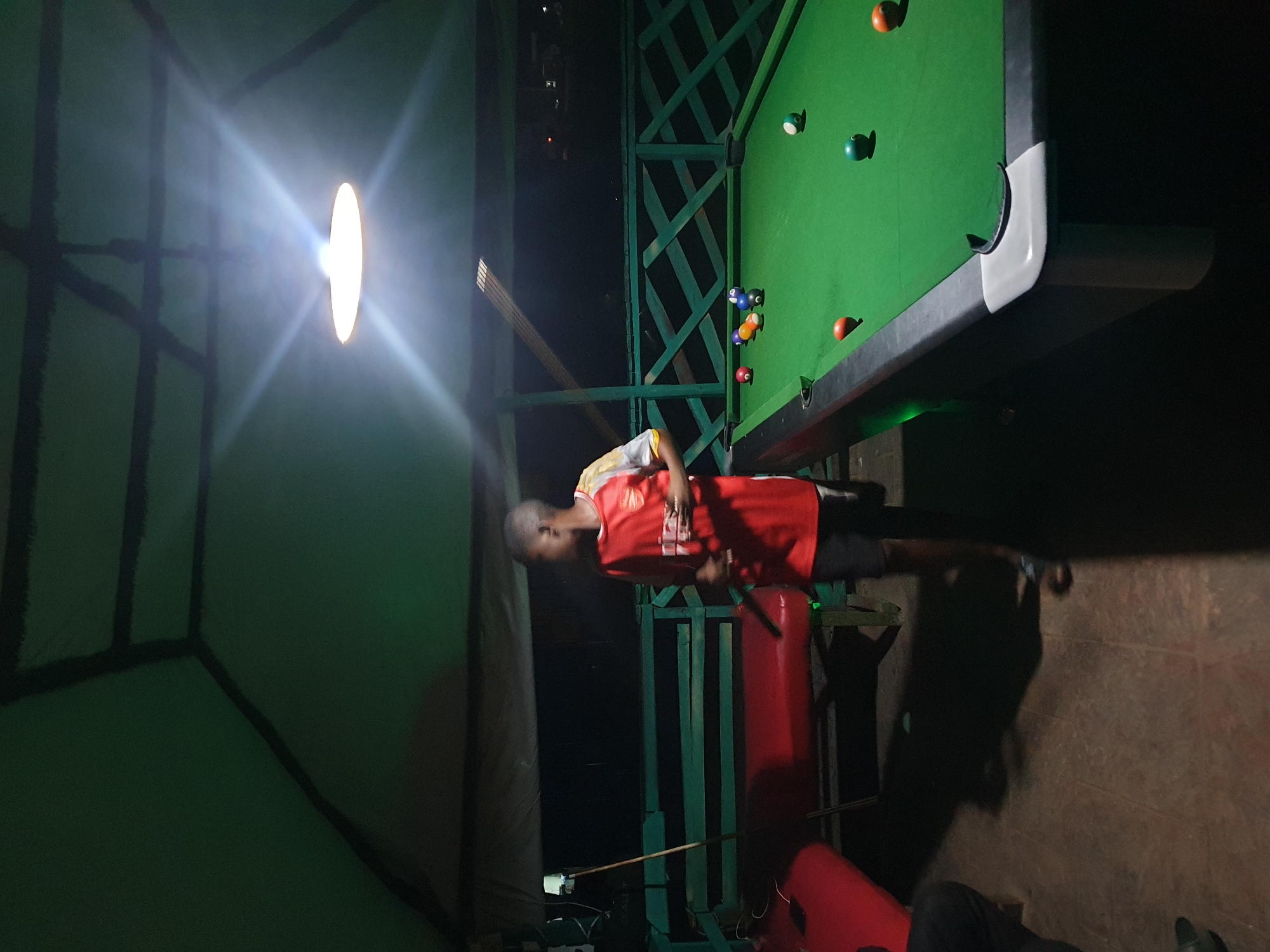 A young Black male wearing a red and yellow sports jersey and dark shorts stands near a pool table under a green tarp roof. He is looking to his right and holding a smartphone. The pool table, which is covered in green felt, has several colored billiard balls on it. To the left of the pool table is a red bench. The scene appears to be an outdoor recreational area, possibly at a bar or private residence, in Lokoja, Nigeria, evidenced by the tropical climate suggested by the open-air setup.