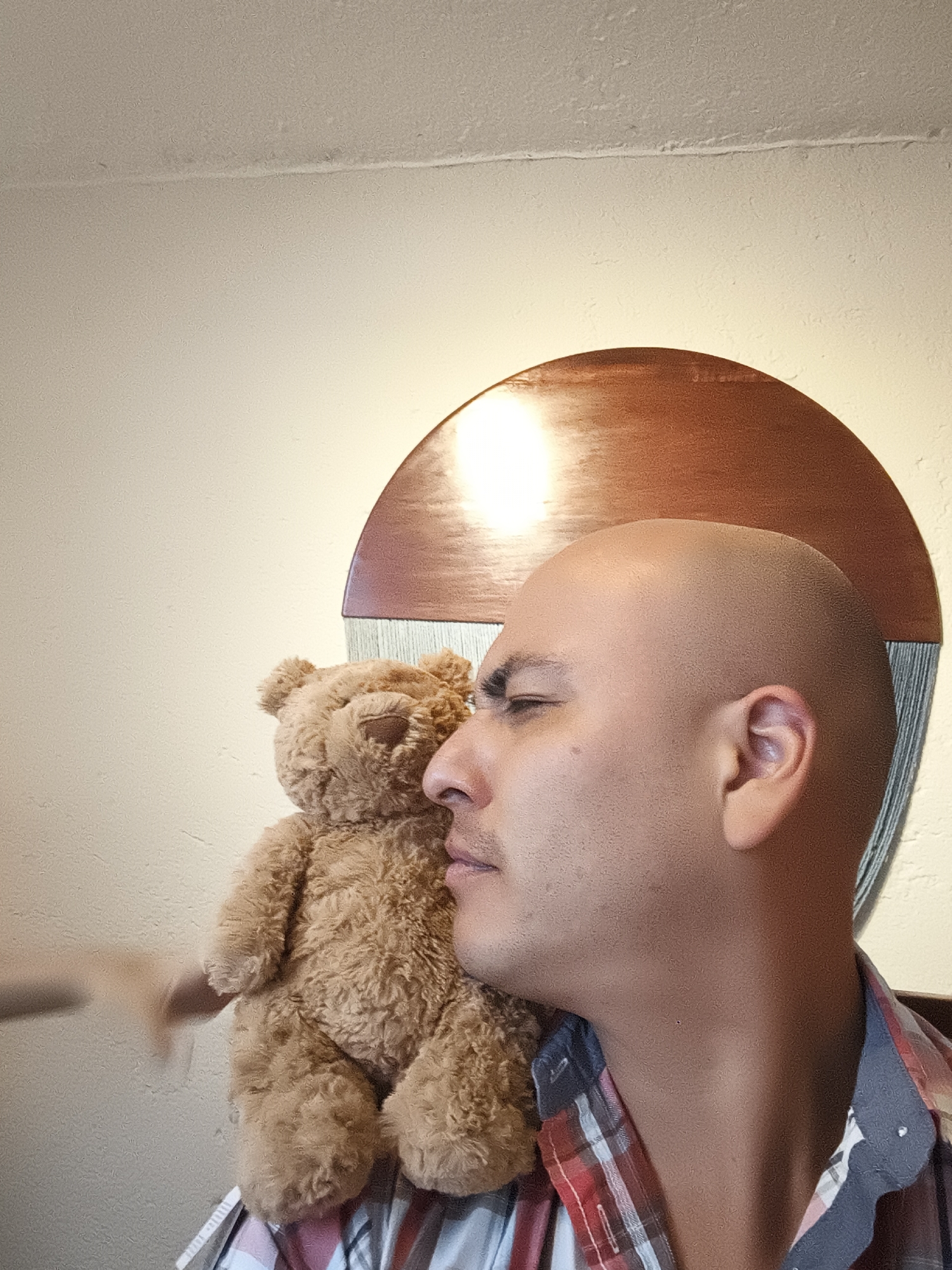 A bald man is hugging a teddy bear, with his cheek against the bear's fur. He is wearing a plaid shirt with a blue collar. Behind the man is a round, wooden headboard with a light shining on it. The background is a light-colored wall. The location appears to be indoors. There is no information to definitively place this image in Mexico City, Mexico.