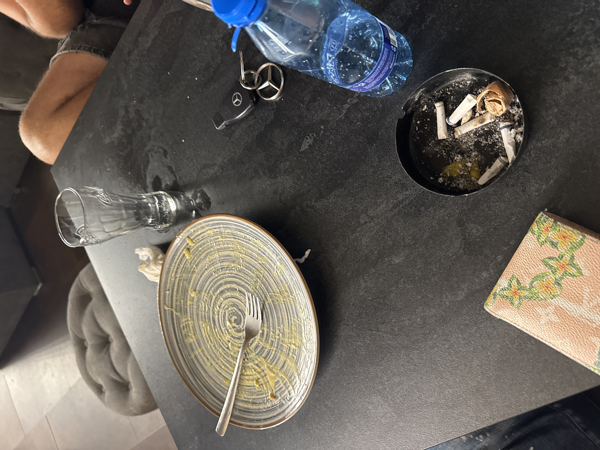 The scene depicts an indoor table setting in Targovishte, Bulgaria. A dark-colored table surface displays various items. An empty plate with a spiral pattern contains yellow food residue and a fork. An empty, ribbed drinking glass and a crumpled white tissue are positioned near the plate. A set of Mercedes-Benz car keys with a fob rests on the table. A partially full blue plastic water bottle, labeled "400 ml," is also present. A black ashtray holds several cigarette butts and ash, including one hand-rolled item. A light-colored wallet or small notebook with a floral pattern is visible in the lower left foreground. A person's leg, clad in shorts and exhibiting hair, is partially visible on the right side of the image. The background includes a tufted cushion, light-colored tiled flooring, and dark furniture. The table surface shows smudges and water rings.