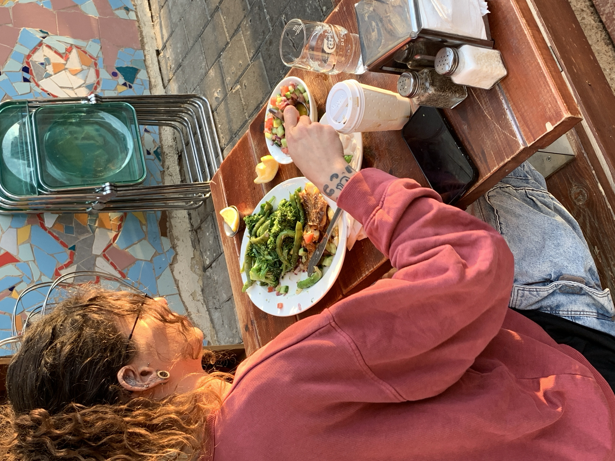 An outdoor dining scene in Tel-Aviv, Israel, captured in warm, late-day sunlight. A person with curly brown hair tied in a ponytail, wearing a maroon long-sleeved sweatshirt and light-wash jeans, is seated at a wooden table, viewed from the back and right side. Their right hand holds a utensil, preparing to scoop food from a small white bowl containing a diced salad of cucumbers, tomatoes, and red beans.
On the table in front of the person is a larger white plate with a partially consumed meal consisting of green vegetables (broccoli, asparagus) and what appears to be grilled protein. Lemon wedges are scattered on the table and plate. Other items on the table include a clear glass with "Carlsberg" branding holding water, a white disposable cup with a lid, a black smartphone placed face down, a salt shaker, a pepper shaker, and a metal napkin dispenser with white napkins. The person's right forearm features a tattoo. Their right ear has multiple piercings, including a stretched earlobe with a gauge.
In the background, a distinctive wall is visible, adorned with a colorful mosaic pattern in shades of blue, pink, red, white, and orange. Leaning against this wall is a stack of four translucent green plastic folding chairs with metal frames. The ground beneath the table is paved with grey interlocking stones or concrete blocks.