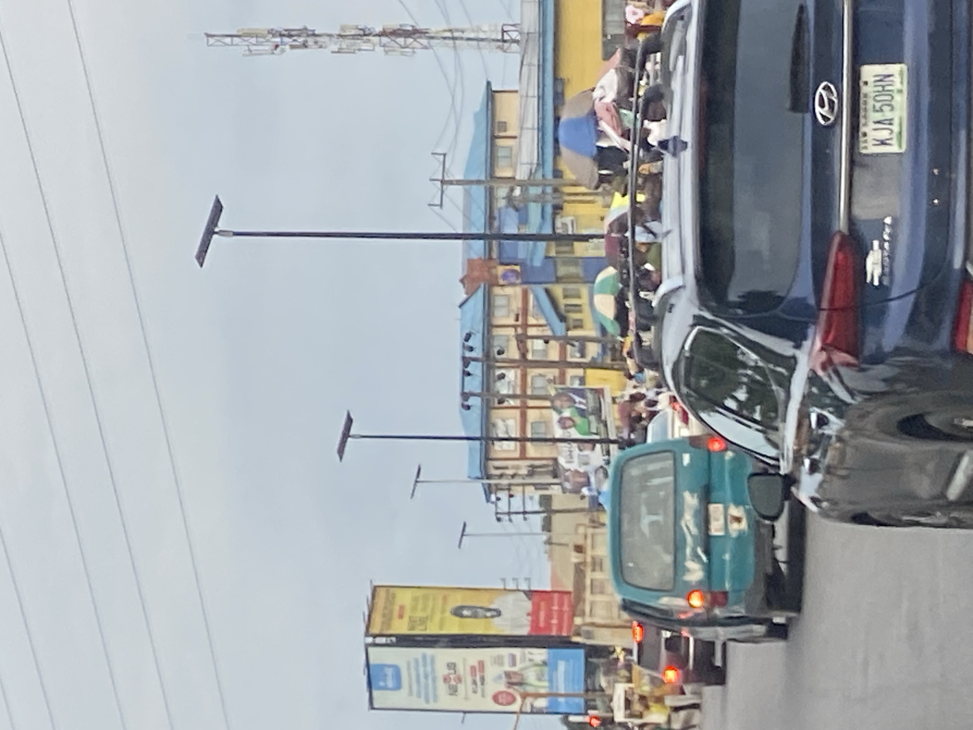 In Ikorodu, Nigeria, a street scene captures a traffic jam under an overcast sky. Tall billboards with advertisements stand prominently on the left side, one featuring a man's portrait and the other an advertisement for "Nexus". A telecommunications tower is visible in the background on the right, along with several buildings with blue and yellow facades. Several vehicles are stopped on the road, including a blue minivan and a dark blue SUV in the foreground. Pedestrians are present on the roadside, some under colorful umbrellas, suggesting a market or vendor area. Solar-powered streetlights are also visible along the road.