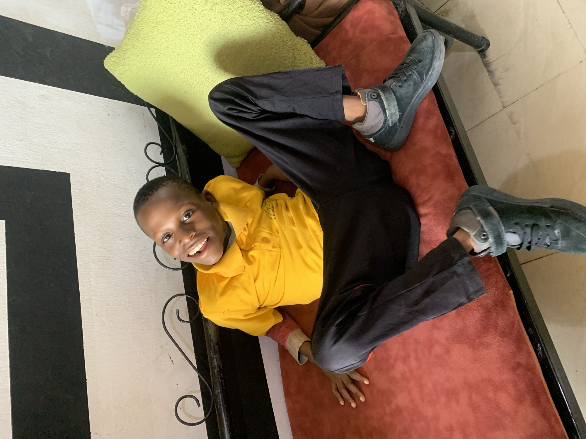 A young boy lies on a reddish-brown sofa with black metal frames, seemingly indoors. He is positioned on his back, with his legs bent upwards, feet directed towards the top of the image. He is wearing a yellow long-sleeved shirt, black trousers, and dark gray sneakers with gray accents. A green pillow is behind his head. He is looking directly at the camera with a smile. The background includes a wall with a black and white striped pattern and beige tiled flooring. This image was taken indoors, likely in a residential space in Jos, Nigeria.