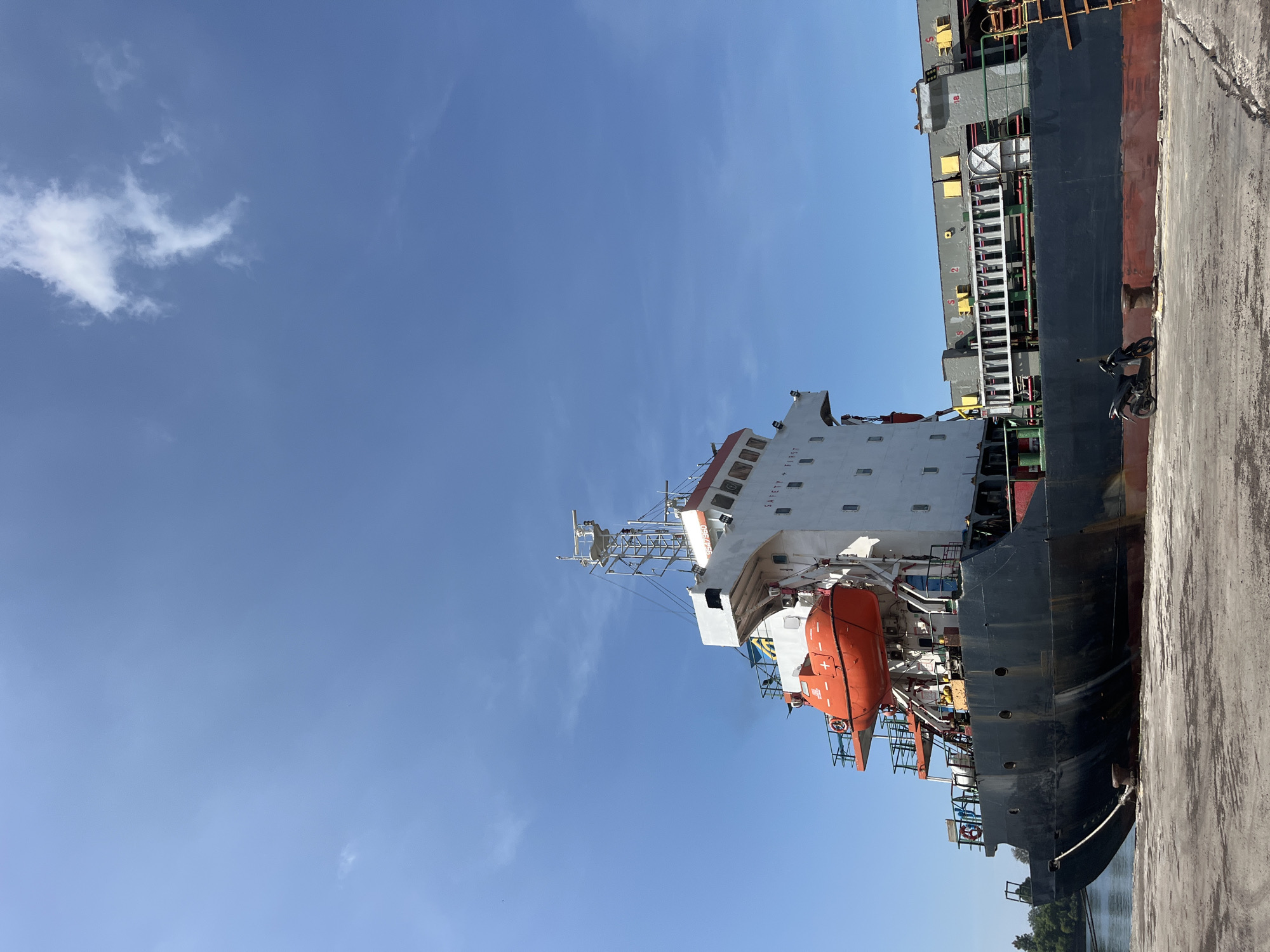 A large cargo ship is docked at a port. The ship is dark colored with a white superstructure. There is an orange lifeboat attached to the side. A gray concrete dock is visible next to the ship. The sky is blue with a few white clouds. The photo was taken in Tambonbaroh, Indonesia.