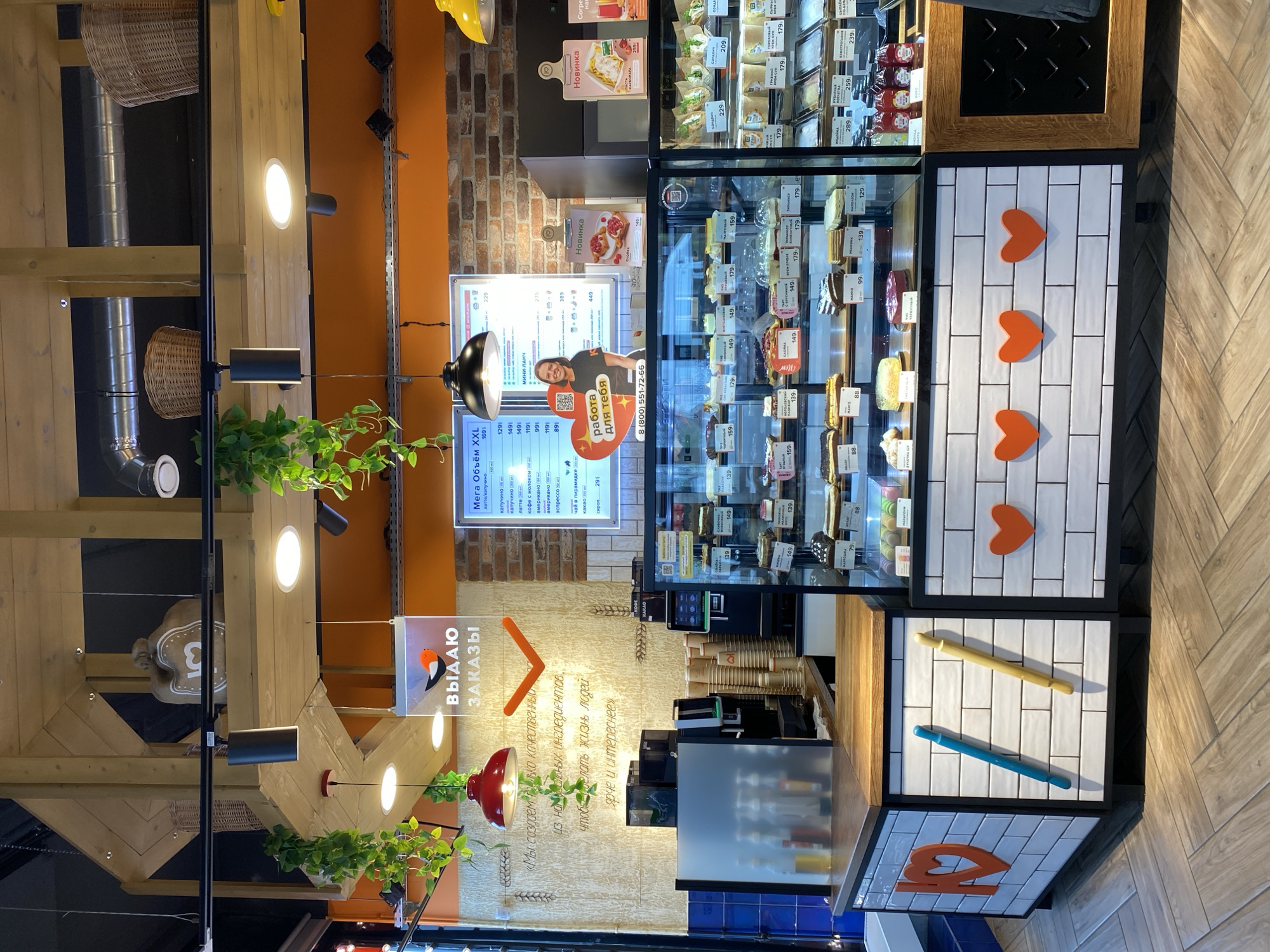 The image captures an indoor scene at a restaurant in Murino, Russia. It shows a counter with a refrigerated display case filled with various desserts. Above the counter is a digital menu board with Russian text and the photo of a person advertising for job. A sign that says "ВЫДАЮ ЗАКАЗЫ", meaning "Orders Issued" with a drawing of a bird and an arrow on it, hangs near the counter. On the front of the counter, there are square tiles with images of hearts, rods and other shapes. The environment includes overhead lighting, wooden structures, and plants decorating the interior. A view of kitchen area with metal pipes are seen on the background.