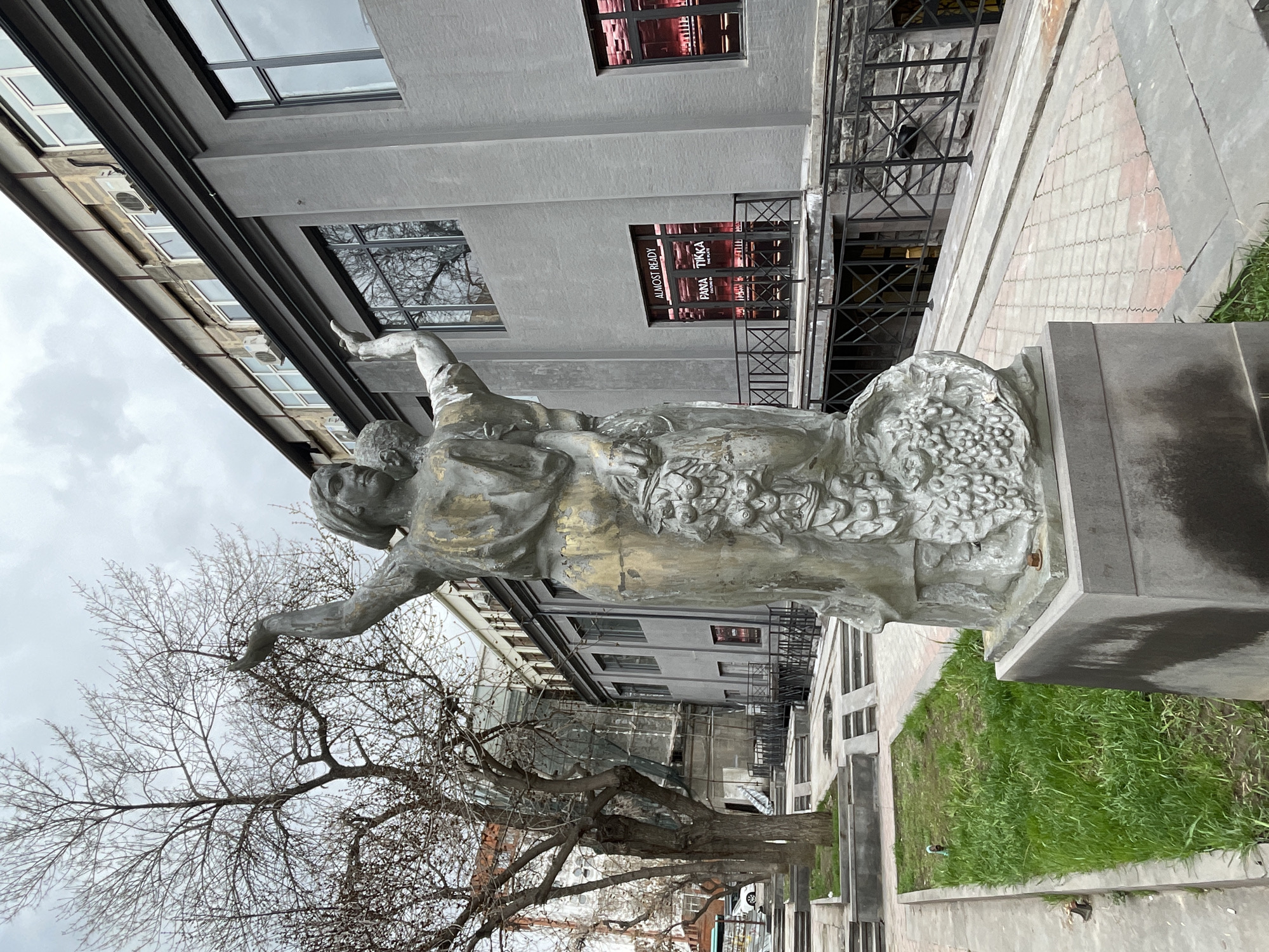 A street-level view depicts a bronze statue of a woman with her arms outstretched, positioned on a concrete plinth in a small patch of grass. The statue is located in Yerevan, Armenia, indicated by the “FANA TIKKA” sign visible in the window of a multi-story building behind the statue. The building is of contemporary design with grey concrete walls and large windows. To the left of the statue is a bare deciduous tree. The sky is overcast. The image appears to have been rotated 90 degrees counterclockwise.