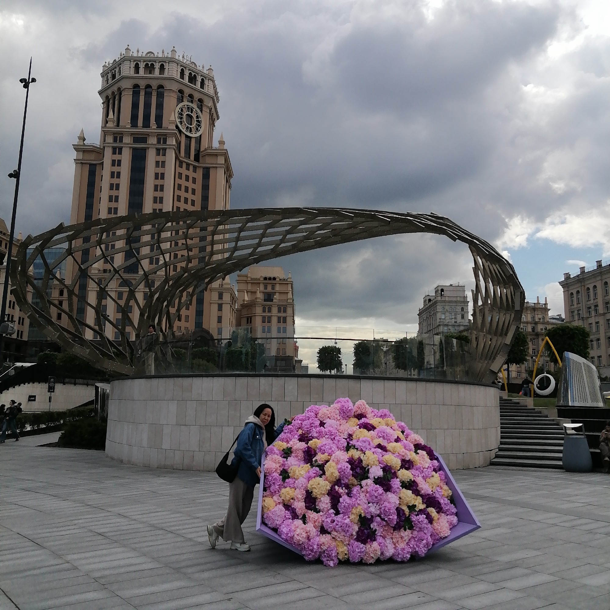 A woman stands next to a large bouquet of pastel-colored flowers in Zaryadye Park, Moscow, Russia. The flowers are arranged in a purple frame, with the woman leaning against the frame with her left hand. She is wearing a denim jacket, beige pants, and white sneakers, and carries a black shoulder bag.
Behind her is a prominent architectural structure, a large, semi-circular metal lattice arch, which serves as a backdrop to the scene. Further in the background is a tall building with a clock tower, indicative of Moscow's urban landscape. To the right, there are stairs leading up to a higher level of the park, with modern art installations and people visible in the distance. The ground is paved with grey stone tiles. The sky above is overcast with dark, stormy clouds.