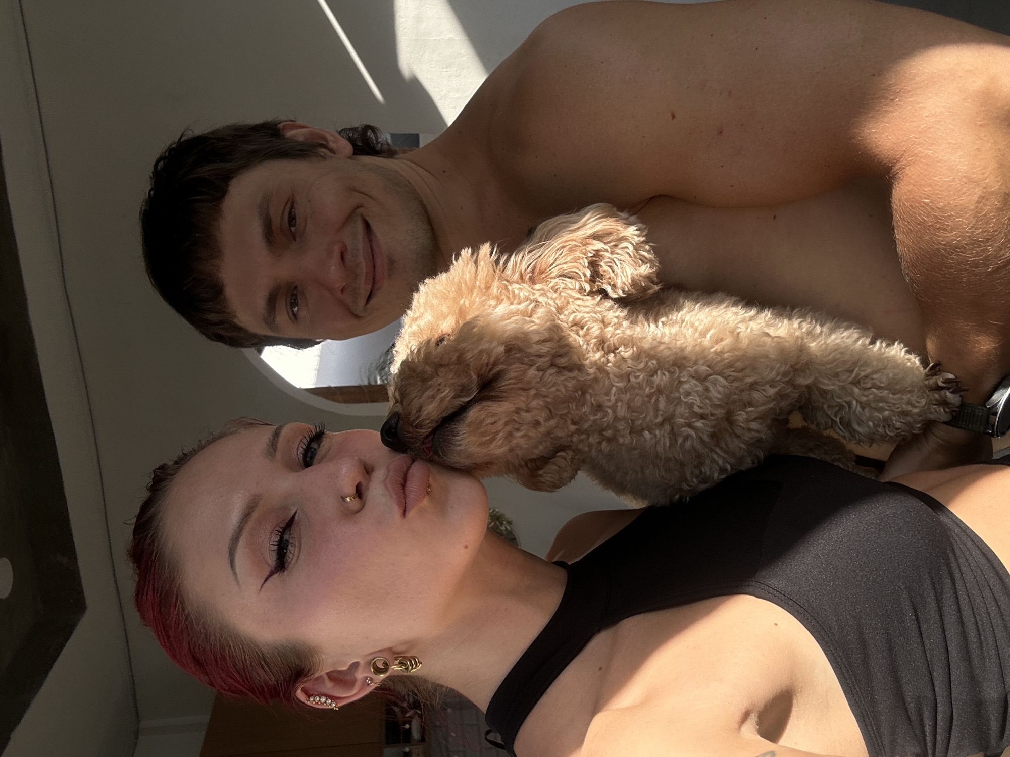 A fair-skinned woman with reddish-purple hair, a nose ring, and multiple ear piercings is positioned in the foreground, wearing a black top. Her lips are pursed, and a small, tan-colored poodle appears to be licking her face. Behind her, a fair-skinned man with short, dark hair is visible. He is shirtless and smiling. The background is indistinct, with shadows and partial views of a ceiling. The location is suggested to be North Kuta, Indonesia.