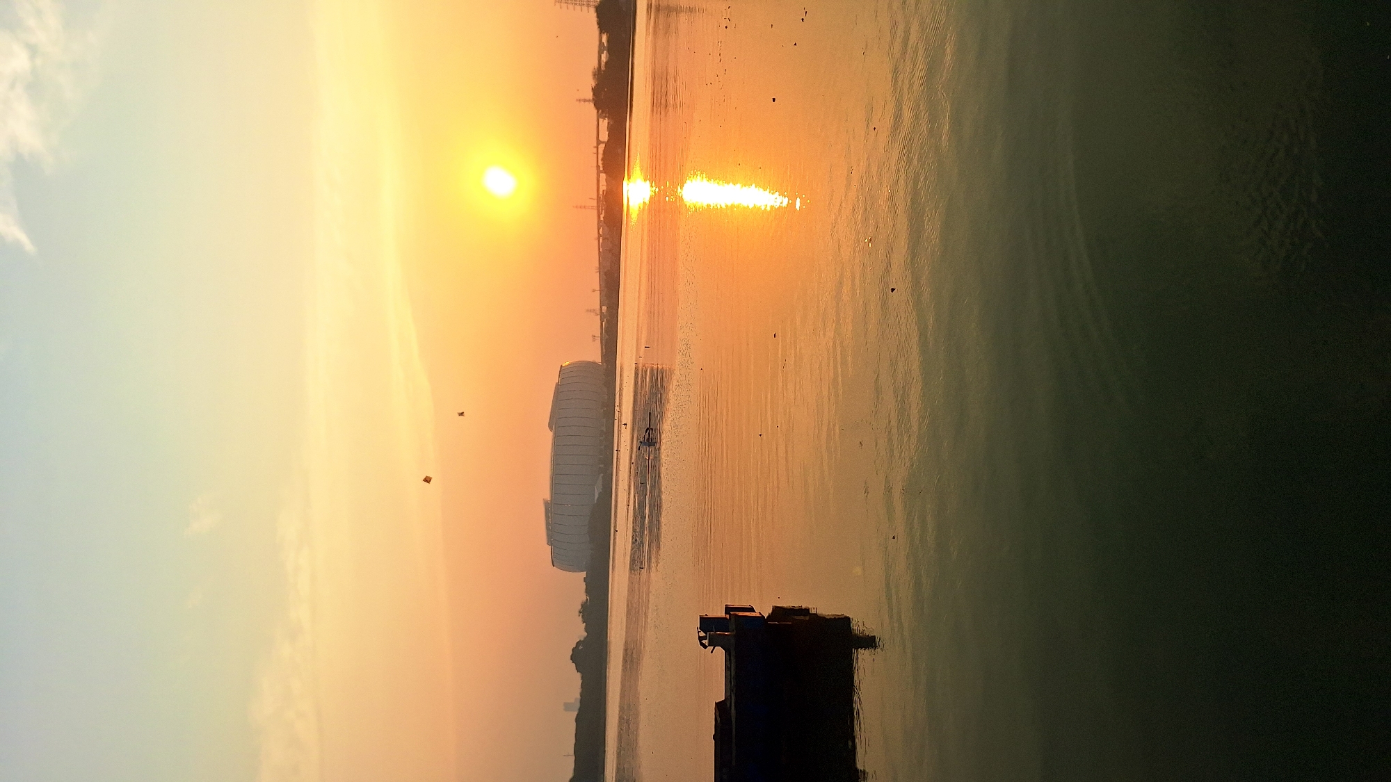 An outdoor, wide shot captures a sunset over the waters of North Jakarta, Indonesia. The sun, a bright orb of yellow, is positioned in the upper right quadrant of the image, casting a warm, orange glow across the sky and reflecting vibrantly on the water's surface. In the middle distance, a large, modern stadium with a distinct oval shape and a striped facade stands prominently against the horizon. Several power lines and some distant buildings are visible on the far shore. A small, dark blue boat is anchored in the foreground on the left, partially submerged in the water. The water itself is calm, with ripples reflecting the sunset colors. A few birds are visible in the sky, and what appear to be two kites are also present. The overall atmosphere is serene and picturesque.