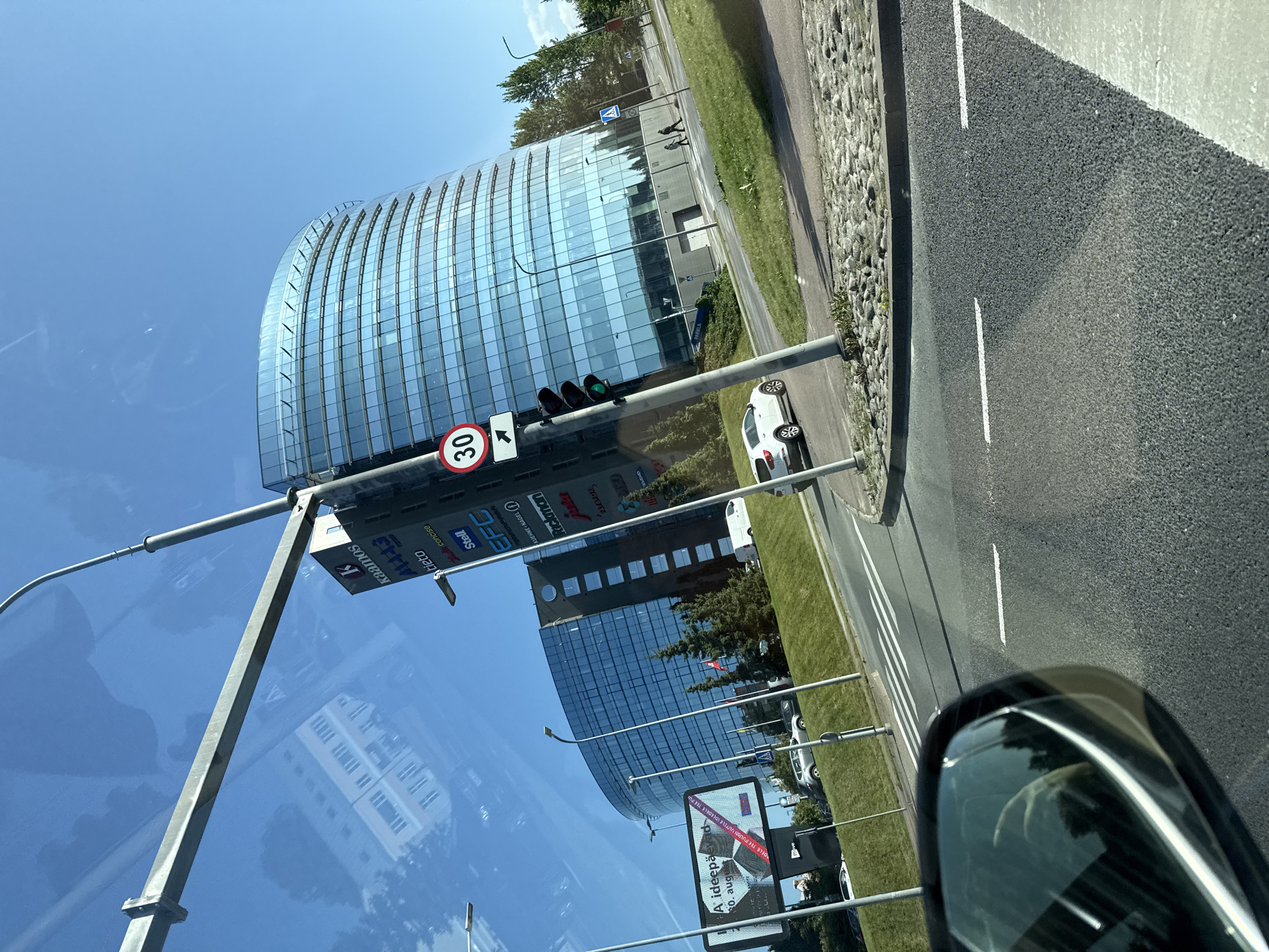 A driver's-side view of a modern, curved glass office building in Tallinn, Estonia, on a sunny day. The building is tall and features numerous horizontal windows reflecting the bright blue sky. A large vertical sign with various company logos, including "kaamos" and "Stell," is visible on a lamppost in front of the building. A 30 km/h speed limit sign is also present on the lamppost, along with a traffic light showing green. A white SUV is parked on the right side of the road, and other vehicles are present in the background. A billboard advertising an event is to the left of the building. The foreground shows a road with white lane markings and a cobblestone curb. The reflection of the sky and nearby buildings is visible on the car's windshield and side mirror. Two people are walking on the sidewalk in the distance.