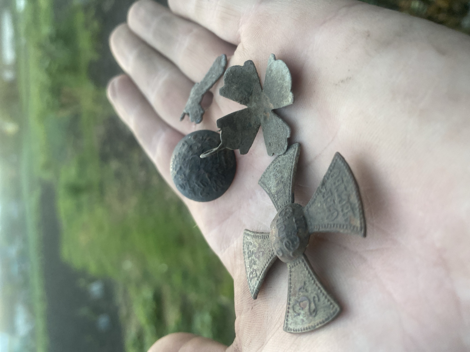 A person's open palm displays four tarnished metal objects. These objects consist of a small, round, possibly hammered button; a stylized four-petal flower; a small unidentified fragment; and a Maltese cross. The Maltese cross appears to have some inscribed text or designs on it. The setting appears to be outdoors, with a blurry background showing some green vegetation and possibly a body of water. There is no indication of the specific location in Ustyvytsia, Ukraine.
