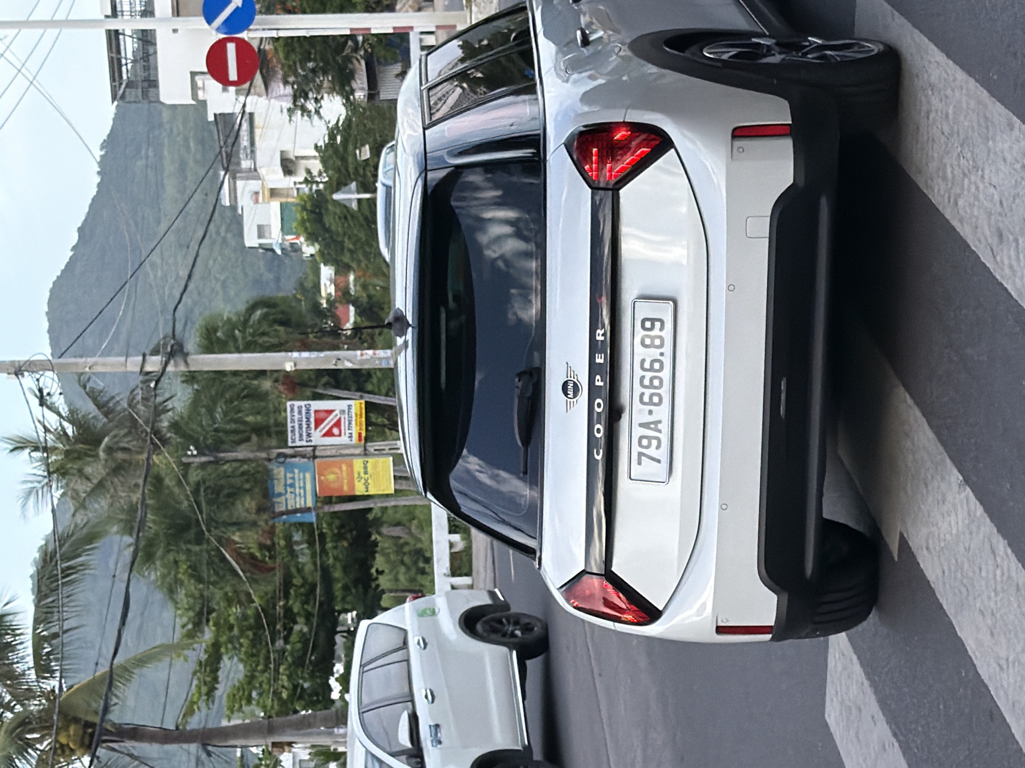 This is a street-level, medium-shot photograph taken in Nha Trang, Vietnam, likely during daylight hours, based on the lighting. The scene features a white Mini Cooper car from the rear, positioned on a pedestrian crossing. The car has a black stripe across the rear with the word "COOPER" in capital letters. Its license plate reads "79A-666.89." To the left of the Mini Cooper, the rear of a white SUV is visible, also on the road. The background shows a street with buildings, lush green trees, and palm trees, indicative of a tropical climate. Several utility poles and overhead wires are present, along with traffic signs, including a red circular sign with a horizontal white bar, signifying "do not enter," and a blue circular sign with a white arrow pointing right. Further in the distance, a verdant, sloping hill or mountain is visible under a hazy sky. The pedestrian crossing has black and white stripes.