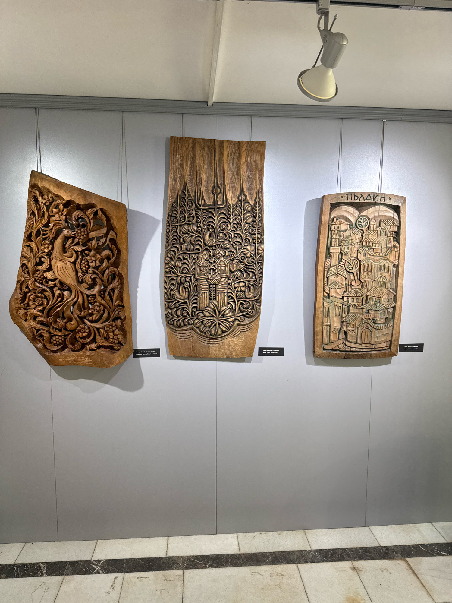 The image depicts three carved wooden panels displayed on a light grey wall, likely in an exhibition or gallery space. The panels are of varying sizes and feature intricate designs. The leftmost panel is irregularly shaped and showcases a bird amidst floral and fruit motifs. The central panel is rectangular and displays a dense, repetitive pattern of leaves and flowers. The rightmost panel is also rectangular and features a detailed carving of a cityscape, with buildings, roofs, and possibly a tree in the center, and the word "ПЛОВДИВ" inscribed at the top. Below each panel are small black plaques, presumably containing descriptive text. A track lighting fixture is visible on the ceiling, illuminating the artworks. The floor beneath the wall is tiled with light-colored squares and a darker inlaid border. The overall setting appears to be a well-lit indoor exhibition space, but no specific location within the city of Pravets, Bulgaria, can be identified from the image alone.