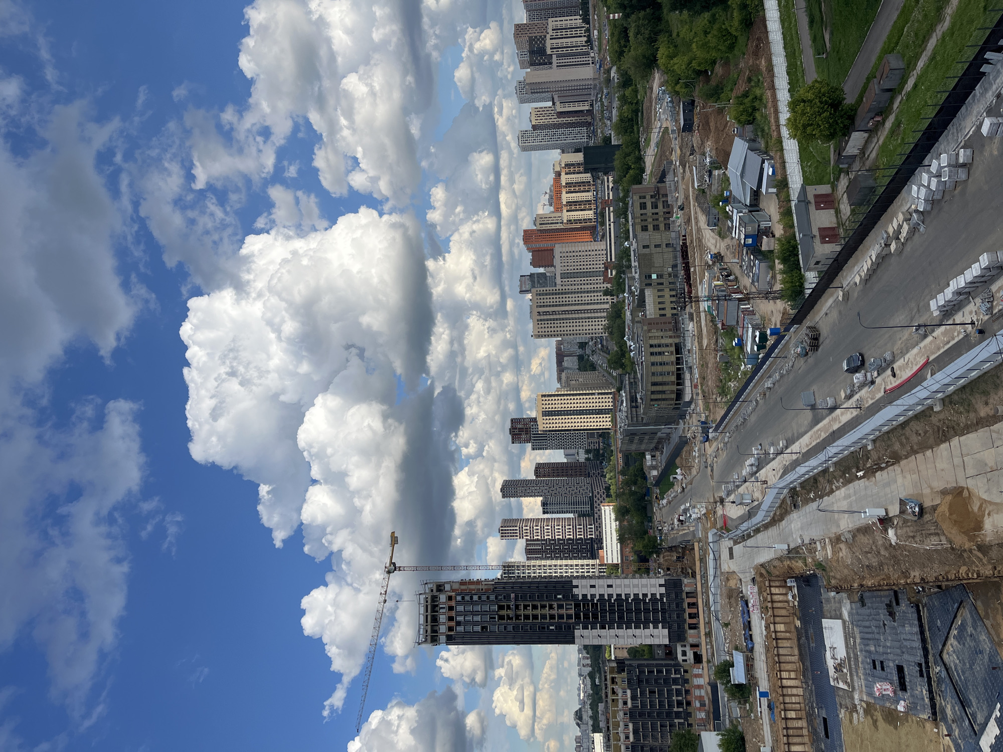 This is an aerial, eye-level, medium shot of a modern urban construction site in Korolyov, Russia, on a bright, overcast day. Dominating the left side of the frame is a tall, multi-story building under construction, with a construction crane extending from its side. In the background, a cluster of modern high-rise apartment buildings, mostly of neutral colors with some accent colors like orange and red, stretches across the horizon. The sky is a vibrant blue, filled with large, fluffy white and gray cumulus clouds. The foreground shows a street under construction, with sidewalks being laid, stacked materials, and temporary fencing. A few vehicles, including a dark-colored car, are visible on the street. The ground is a mix of dirt, concrete, and construction materials. The surrounding area is lush with green trees, indicating a possible park or residential area adjacent to the construction. The overall scene depicts ongoing urban development in a growing city.