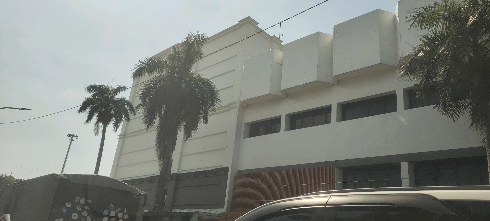 A wide-angle outdoor shot depicts a contemporary white building with geometric architectural features and palm trees in front of it. The building has a light-colored facade with horizontal lines and protruding cubical elements on the upper levels. Rows of dark-tinted windows are visible on the middle and lower floors. To the left of the building, a tall, slender street lamp and power lines stretch across the frame, with a portion of a gray, covered structure partially visible in the foreground. Two palm trees, one taller than the other, are situated in front of the building. The sky is overcast, suggesting a daytime scene. The image was taken from a low vantage point, possibly from a vehicle, looking up at the building. The scene is characteristic of a commercial or institutional area in a city, and the presence of palm trees indicates a tropical or subtropical climate, consistent with Semarang, Indonesia.