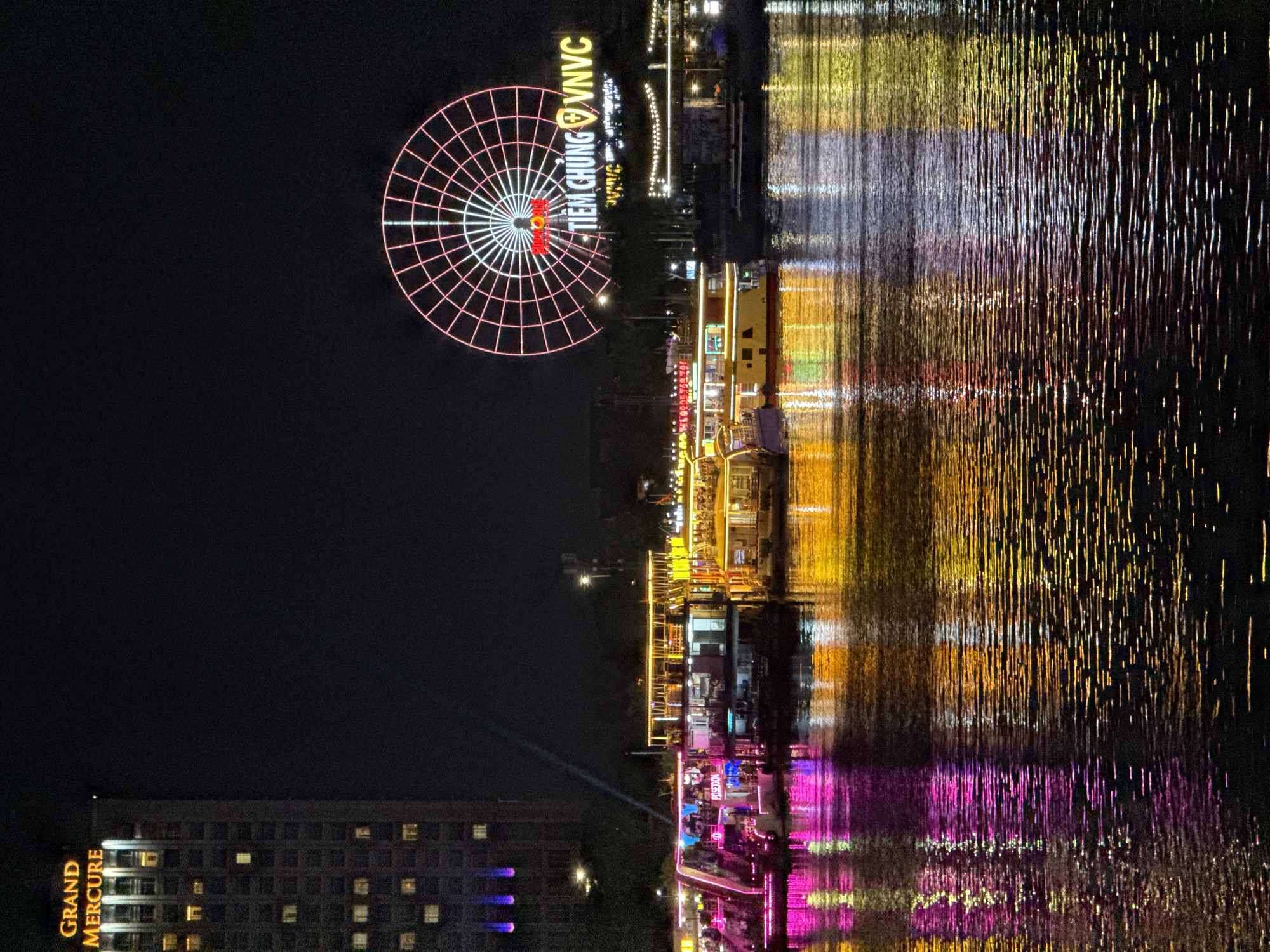 This media file presents a vibrant nighttime cityscape along a river in Đà Nẵng, Vietnam. The sky is dark, indicating a clear night, and the scene is awash with a multitude of artificial lights, which create striking, elongated reflections on the water's surface.
On the left side of the frame, a tall building stands, identified by the vertically oriented golden text "GRAND MERCURE" along its side. Its numerous windows glow with warm light.
Dominating the center-left of the image is a large, brightly illuminated Ferris wheel. Its circular structure is outlined in red lights, with a radial pattern of white lights converging towards its hub. Visible text on the Ferris wheel includes "TIEM CHUNG VNVC" and, partially, "PHƯƠNG ĐÔNG" in red on its central white section.
To the right, a wide river or waterway stretches, featuring several multi-story boats or dinner cruises. These vessels are extensively decorated with colorful lights, primarily in shades of yellow, orange, and magenta, contributing to a festive atmosphere. The reflections of these lights, along with those from the illuminated riverbanks and distant structures, stretch across the water in wavy, vertical streaks of vivid color, contrasting sharply with the dark water.
The overall scene depicts a lively urban waterfront at night, suggesting a popular area for tourism, leisure, and dining.