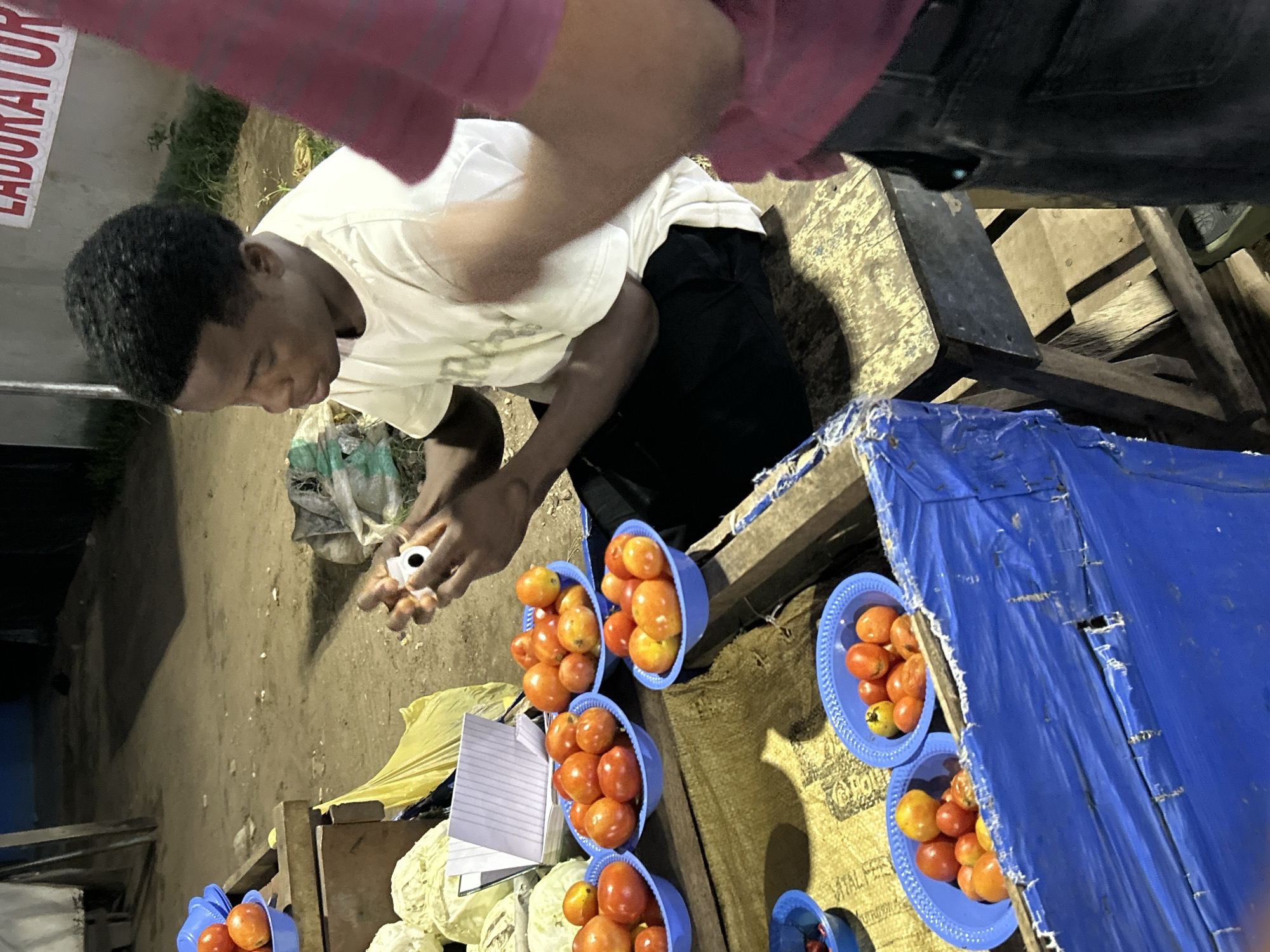 An outdoor market stall in Ihiagwa, Nigeria, at night. A young Black man wearing a white t-shirt and dark pants sits on a wooden stool, holding a small white object with a black circle in his hands. In front of him are several blue plastic bowls filled with tomatoes of varying sizes and shades of red and orange, some of which appear to be cherry tomatoes. Behind the tomatoes are some heads of cabbage and a yellow plastic bag. In the background, a red sign with white lettering reads "LABURATOR." Another person, partially visible on the right, wears a pink shirt and dark pants. The ground is dirt and debris.