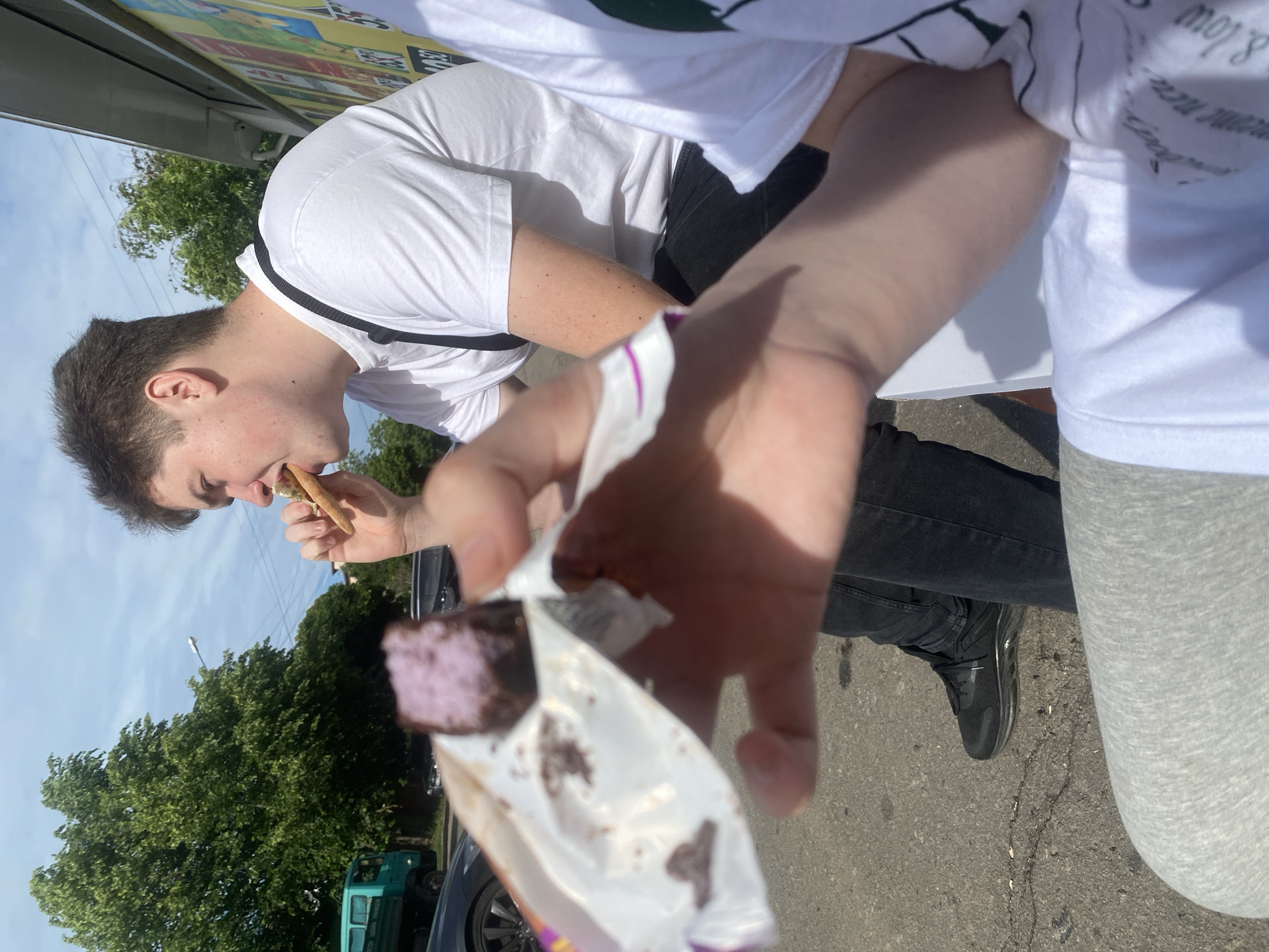 A photo depicts two people outdoors. One individual, wearing a white t-shirt with black straps, is eating a long, thin snack. The second individual is holding an open package containing what appears to be a square-shaped dessert with a purple filling. The foreground is filled with the second individual's arm and hand. The setting seems to be a city street with cars, trees, and buildings in the background. A turquoise truck is partially visible. The location is Poltava, Ukraine.