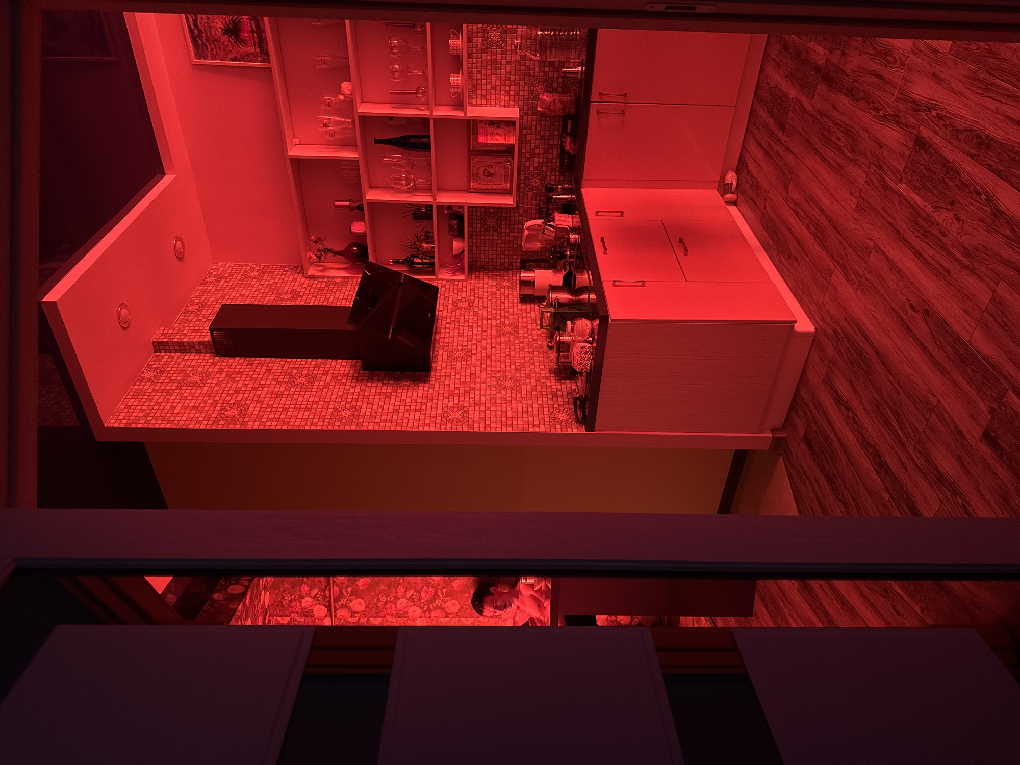 The image depicts a kitchen interior, viewed from a slightly elevated angle, with a red filter applied to the entire scene. Visible elements include white kitchen cabinets, a tiled backsplash, what appears to be a kitchen island with an attached cooking appliance, possibly a grill or stovetop, and tiled flooring. Wooden planks make up the flooring on the right side of the image. The lighting is dim due to the red filter. In the lower center of the image, a person is visible in what seems to be a reflection, suggesting a reflective surface or doorway. The architecture suggests a modern design. Based on the style and content, the location of the image may be Sochi, Russia.