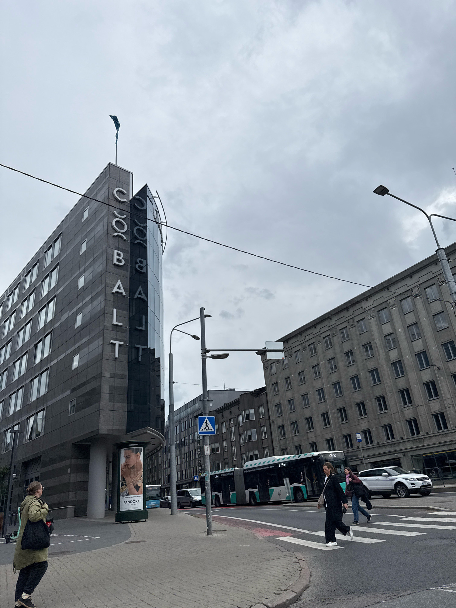 The image shows a street scene in Tallinn, Estonia, on a cloudy day. A modern, angular building with the word "COBALTT" vertically displayed on its side in white lettering stands prominently on the left. The building's facade is made of dark gray stone and glass. A blue flag flies from a pole on the building's roof.
In front of the "COBALTT" building, a large cylindrical billboard displays an advertisement for Pandora jewelry, featuring a tattooed male model.
Across the street, a gray, multi-story building with many windows exhibits a more traditional architectural style.
A public transportation bus with a green and white color scheme is visible in the middle ground, moving from left to right. A white SUV is also seen on the right side of the street, driving in the same direction as the bus.
Several pedestrians are present:
On the left sidewalk, a person wearing a green coat and carrying a black bag walks away from the camera.
In the crosswalk, two individuals are crossing the street from right to left. One person, dressed in black, is in the foreground, while another, wearing a dark jacket and pink scarf, is behind them.
Streetlights and traffic signals are present, indicating a busy urban environment. The road has clear lane markings, including a pedestrian crosswalk. The sky is overcast with a light gray cloud cover. The scene appears to be a commercial or business district of the city.