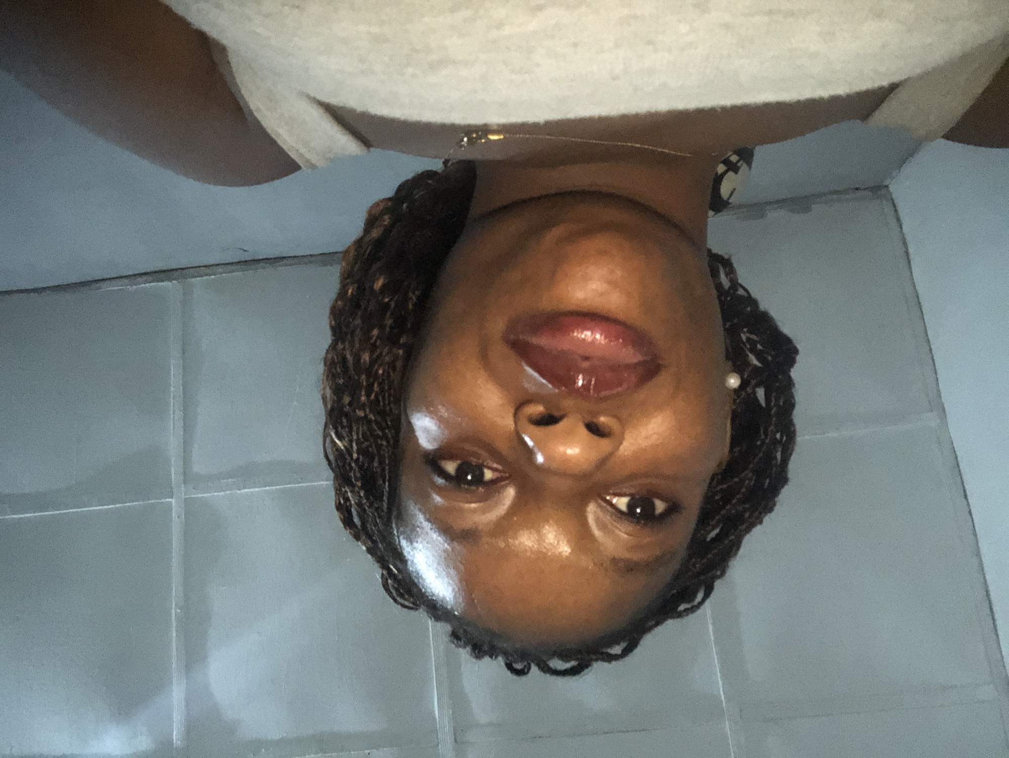 The image shows a woman with dark skin and braided hair, wearing a white tank top and a pearl earring. She is wearing a necklace. The background appears to be a tiled wall. The location is Kashere, Nigeria.