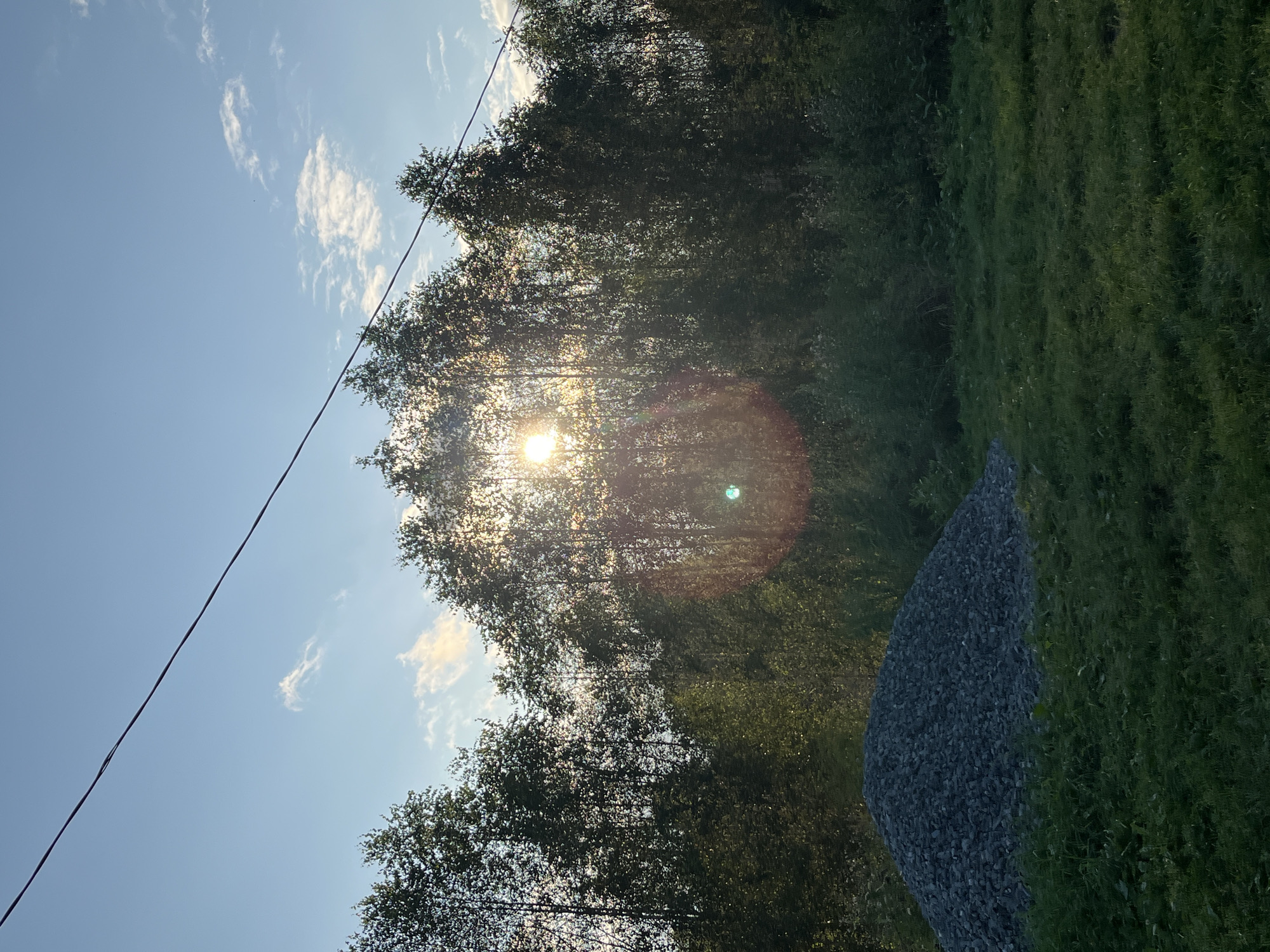 The image captures a natural outdoor scene under a bright blue sky with scattered white clouds. A thick line of trees forms a dense backdrop, partially obscuring the setting sun which creates a lens flare effect with a reddish circular glow. In the foreground, to the left, is a large conical pile of gray gravel. The ground in front of the gravel pile is covered in green grass. A single dark utility wire stretches diagonally across the upper portion of the frame from the top left to the top right. The scene is devoid of any human presence or activity, depicting a still, rural-like environment. The specific location within the city of Nizhny Tagil, Russia, is not discernible from the visual information presented in the image.