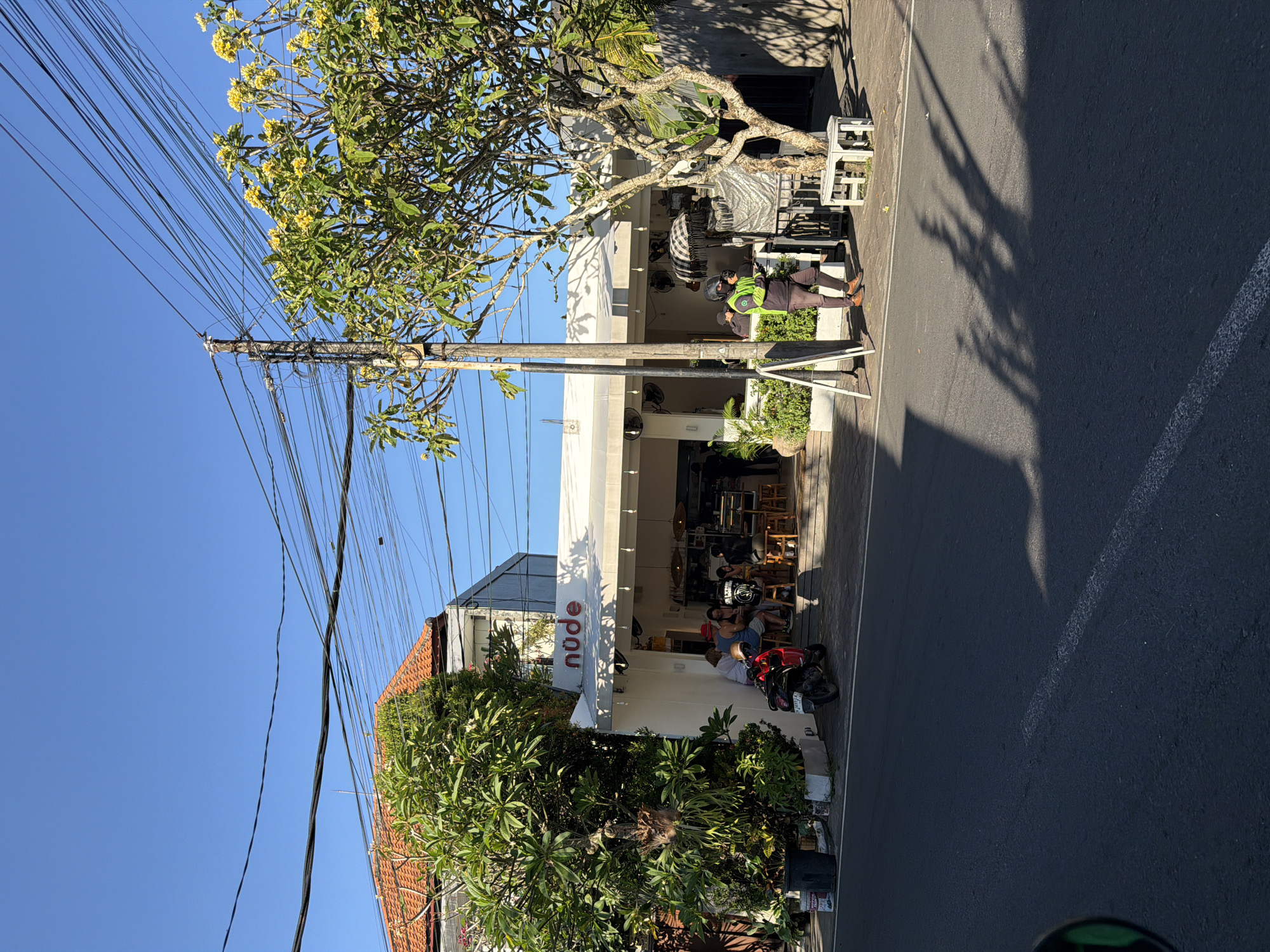 The image captures a street scene in Mengwi, Indonesia, featuring the "nude" cafe prominently. Patrons sit inside the cafe, and motorbikes are parked nearby on the roadside. A utility pole with numerous power lines dominates the left side of the frame, partially obscured by a tree with yellow flowers. Across the street, a person in a green vest is near a building with outdoor seating. Shadows stretch across the paved road, suggesting strong sunlight.